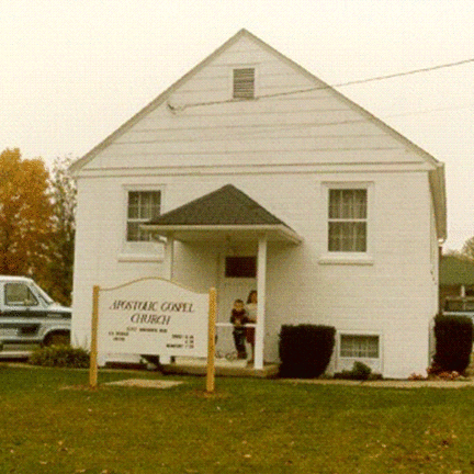 A vintage photo of a white two-story church with a sign in front. There are two people standing near the entrance, and a van is parked nearby. The scene is set on a grassy area with some bushes and trees in the background.