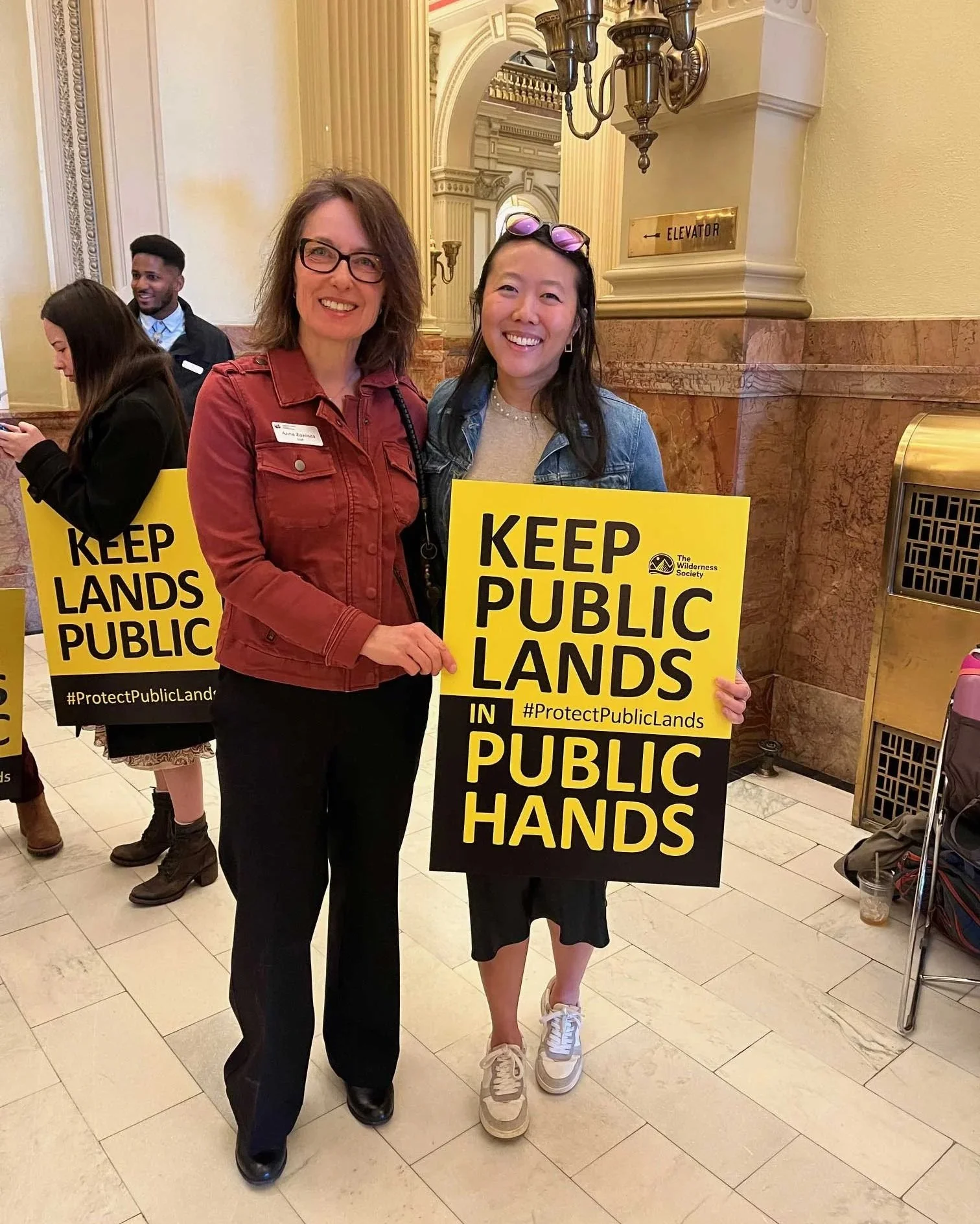 Two people standing inside a formal lobby holding signs that say Keep Public Lands in Public Hands