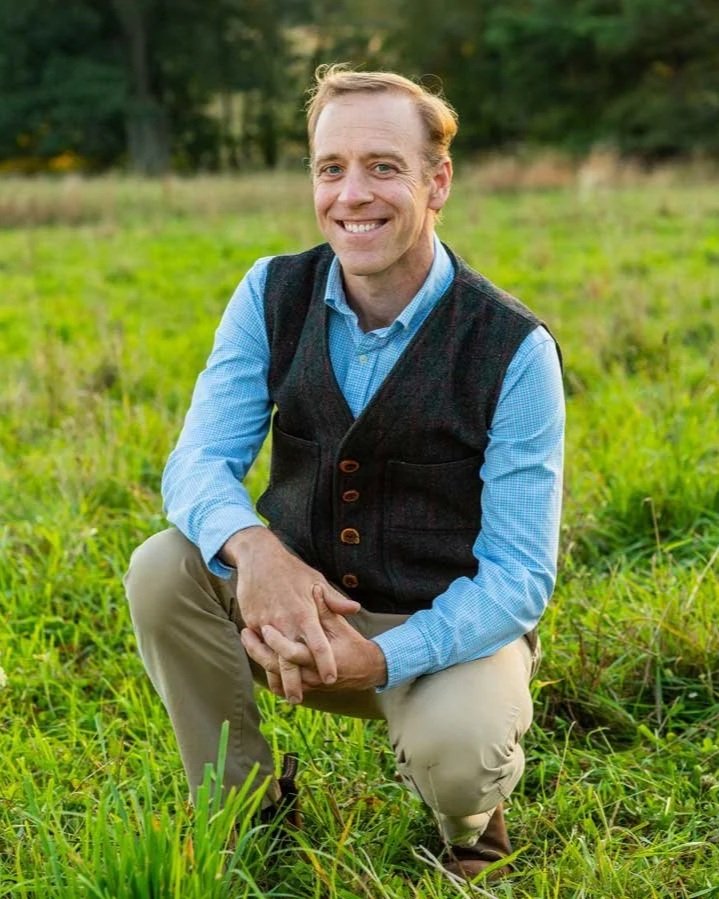 Gregory Horner wearing a blue shirt, dark vest, and khaki pants crouches in a grassy outdoor field with trees in the background.