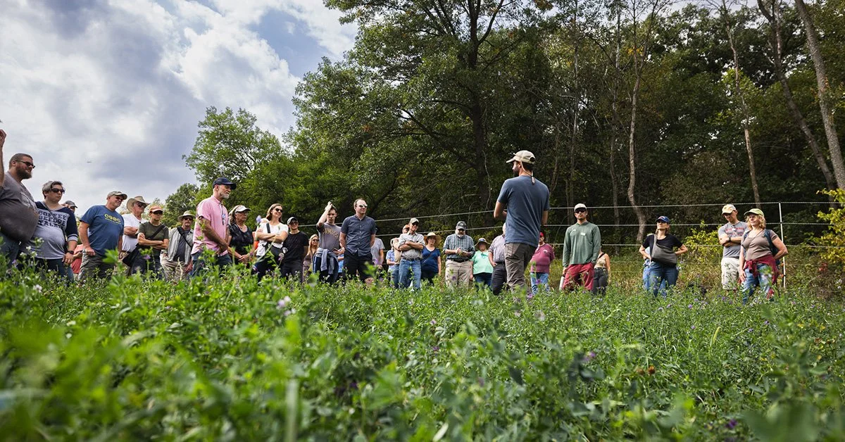 a group of people standing outside in a wild field looking at a person who is speaking to them.