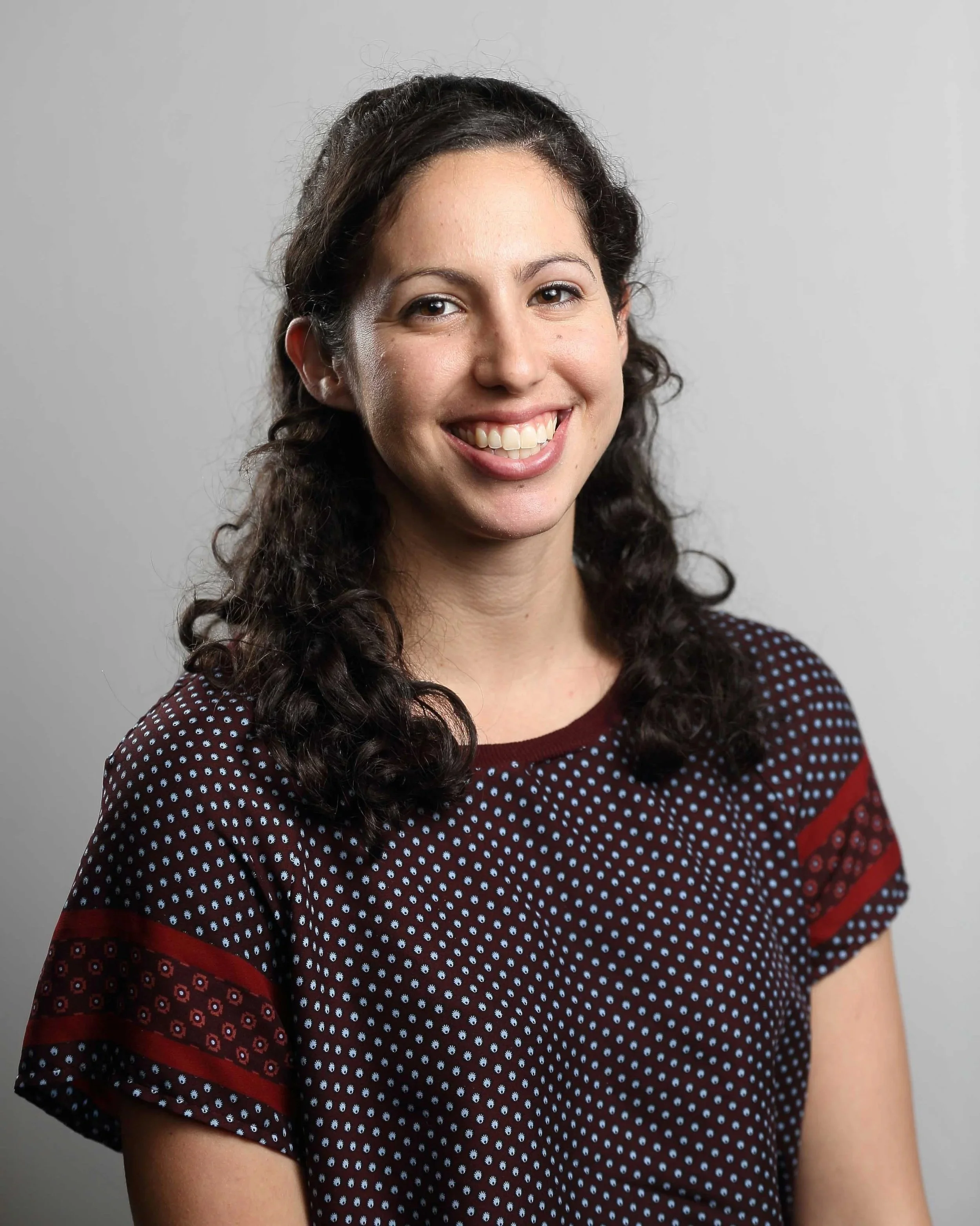 Lucy Winikoff with dark curly hair, smiling, wearing a patterned burgundy top with red and blue accents, against a plain gray background.