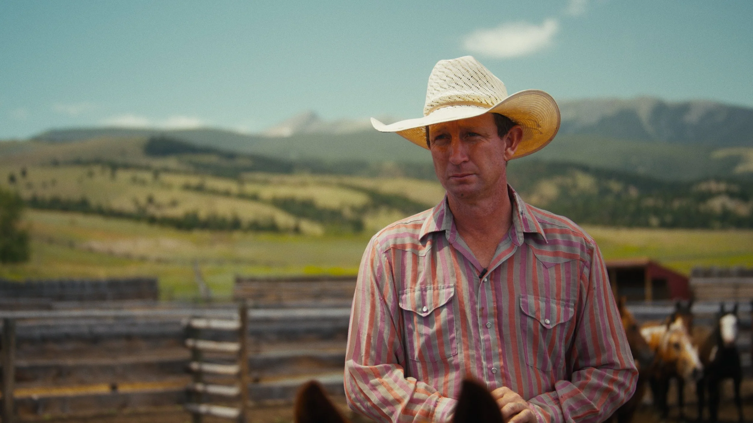 A man wearing a striped shirt and a straw cowboy hat standing outdoors on a farm with hills in the background.