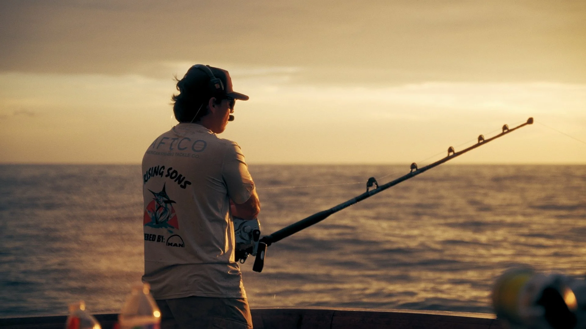 Man fishing at sunset on a boat, holding a rod and looking out over the water.