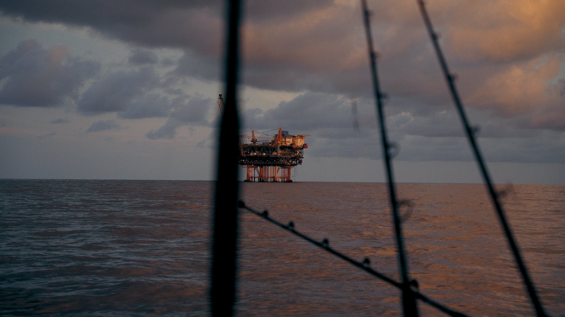 Offshore oil rig in the ocean during sunset, partially obstructed by the image of a boat's rigging.