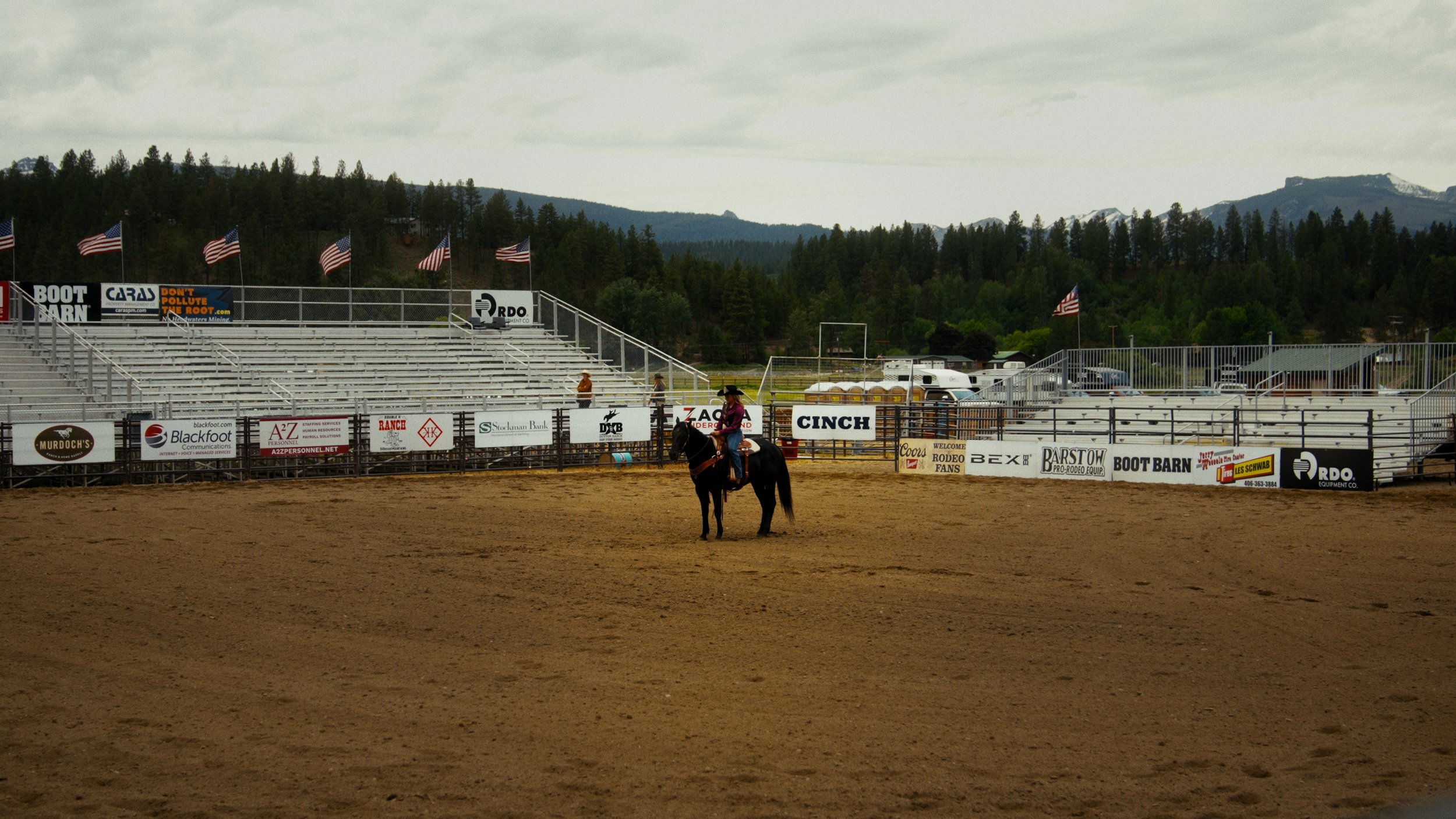 A person riding a horse in an empty rodeo arena with empty bleachers and American flags on the fence, mountains and cloudy sky in the background.