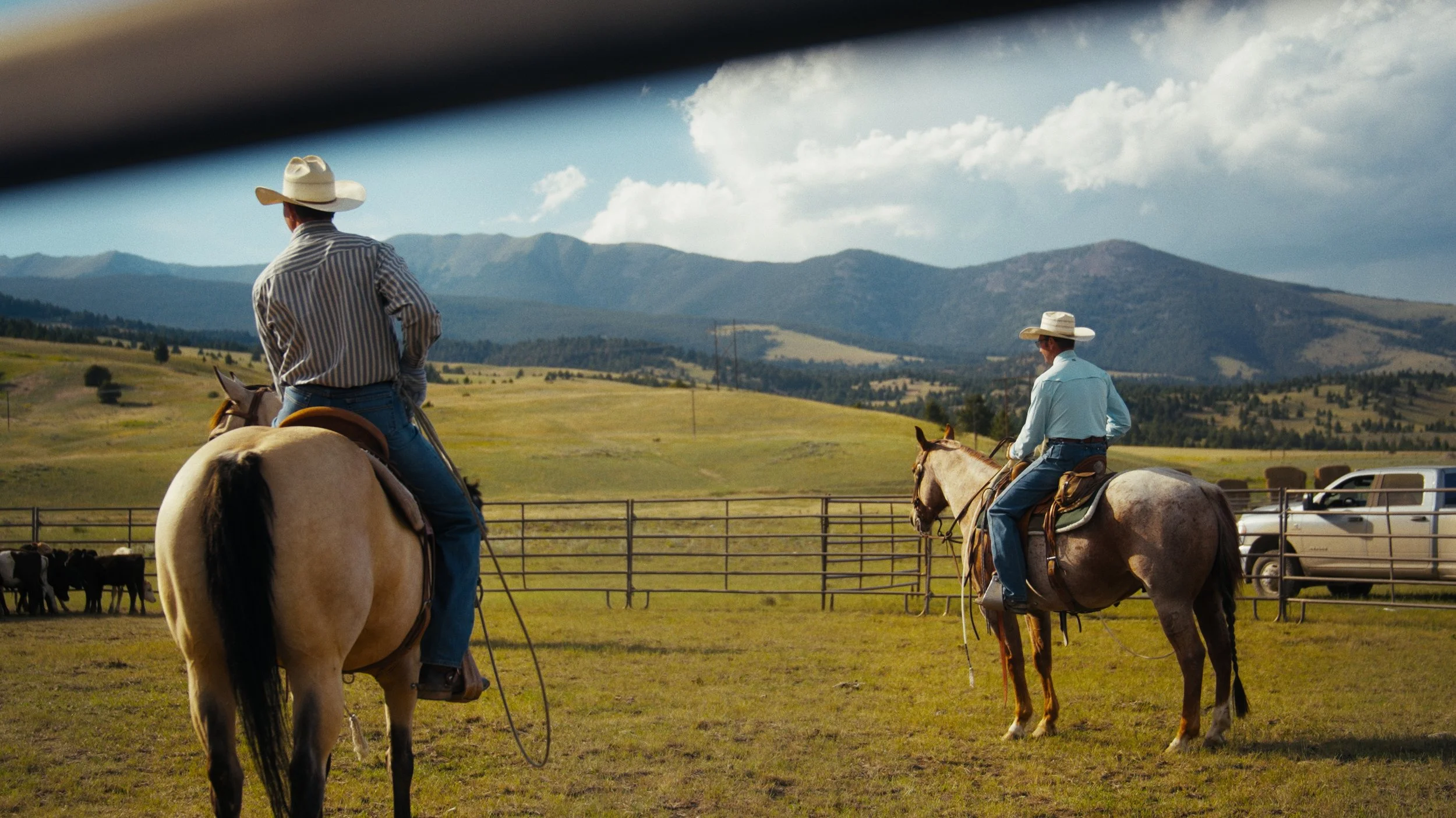 Two cowboys riding horses in a fenced field with mountains in the background under a partly cloudy sky.