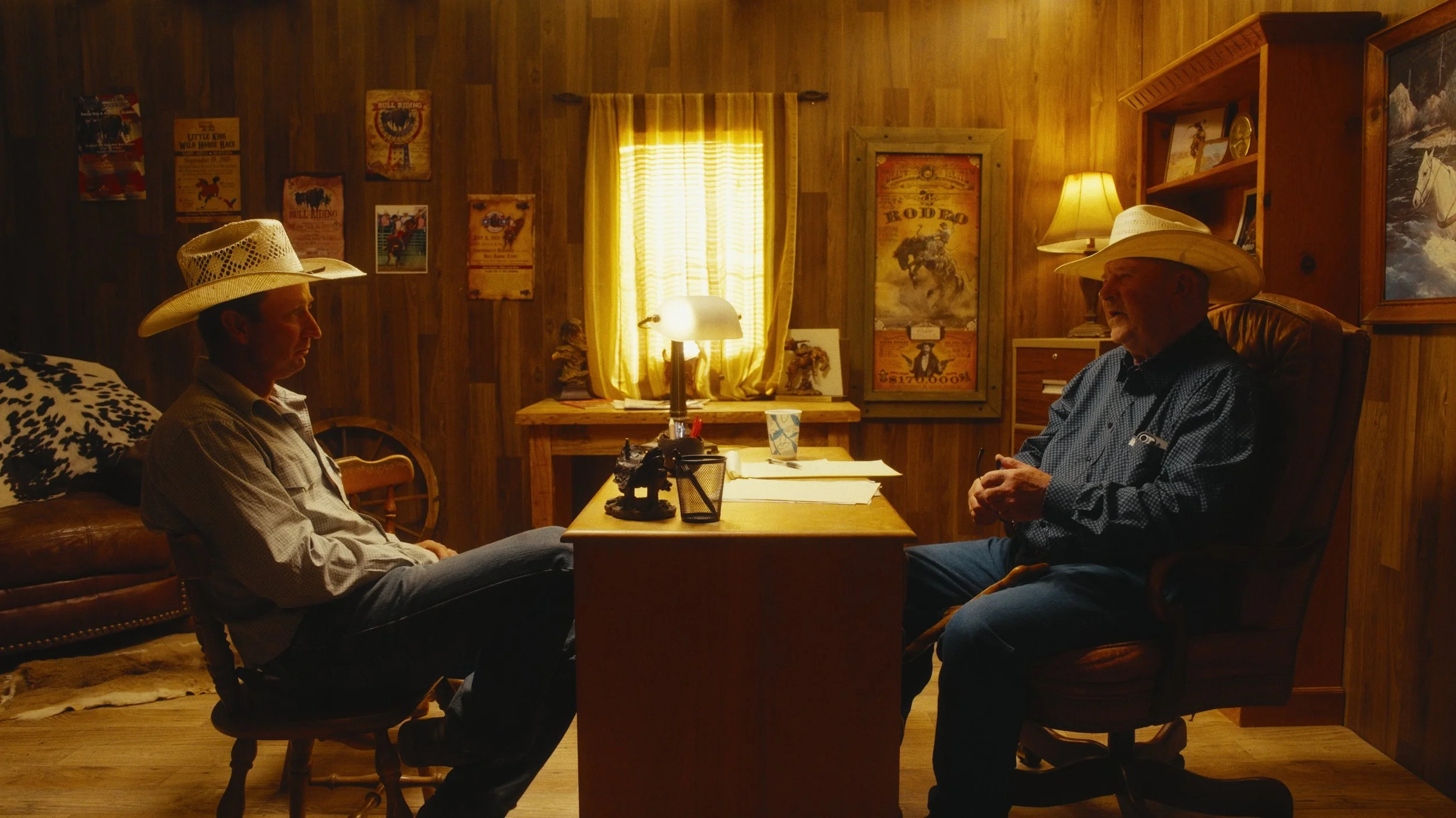 Two men sitting in a wood-paneled room, facing each other, wearing cowboy hats. One is on a leather couch, and the other is in an armchair. The room features posters on the wall, a window with yellow curtains, a desk with papers and a lamp, and various decorations including a framed picture of a horse race.