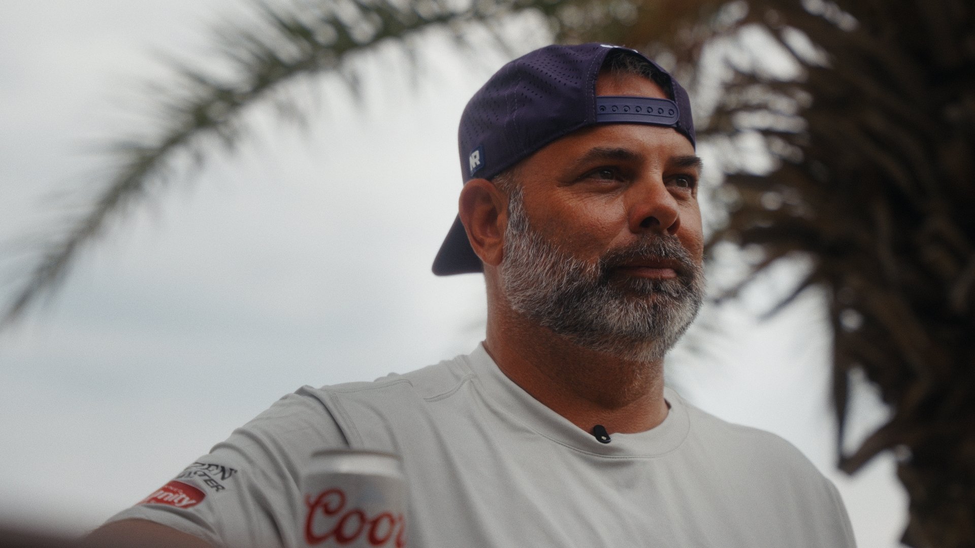 A man with a gray beard and mustache wearing a navy baseball cap backwards and a white t-shirt outdoors with palm trees in the background.