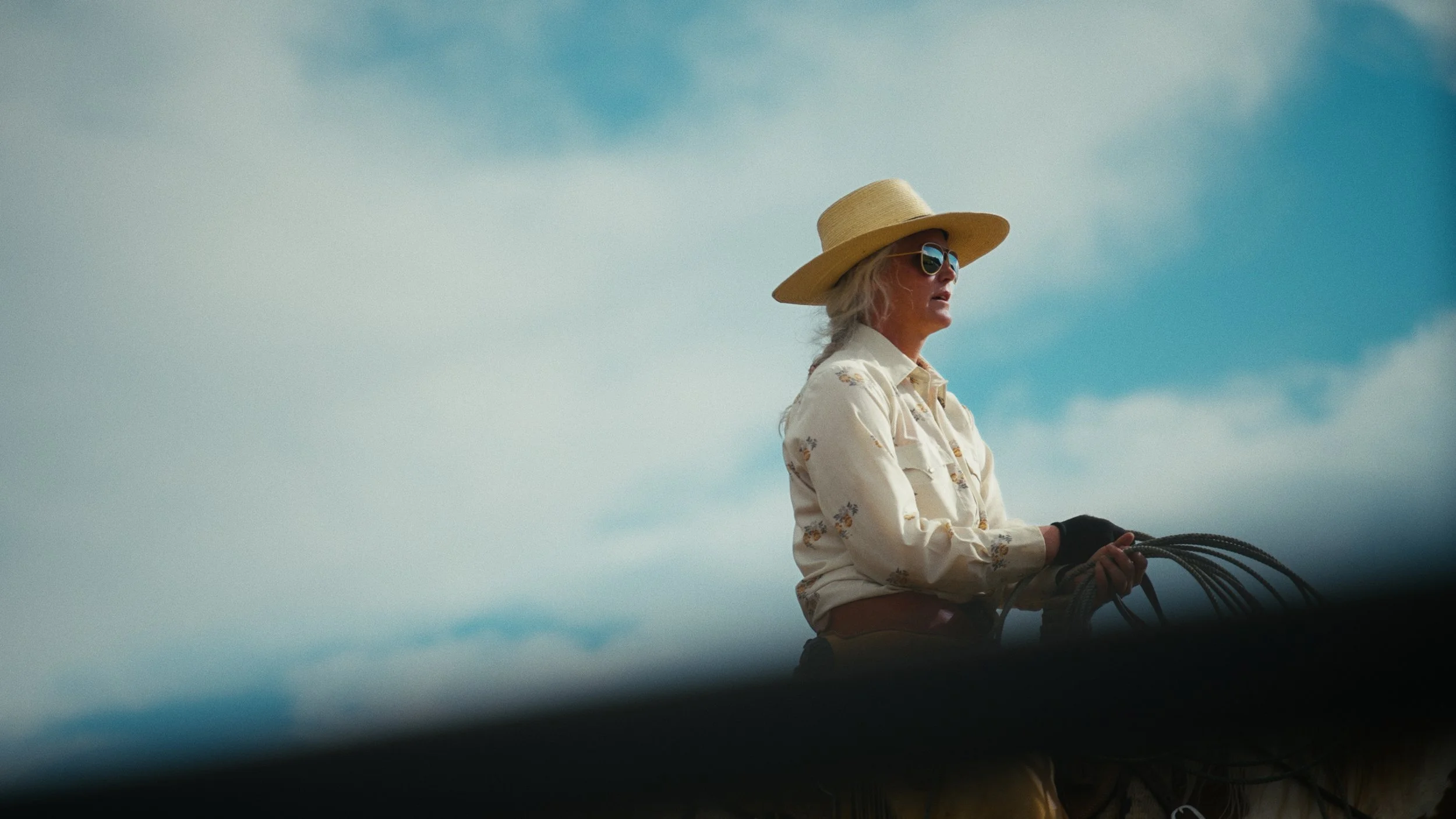 Older woman wearing a wide-brimmed hat and sunglasses, riding a horse against a cloudy sky.