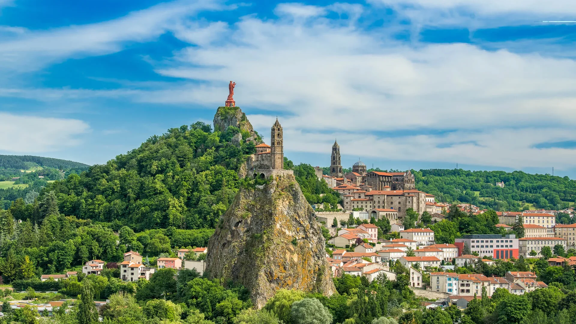 Vue pittoresque d'une ville à flanc de colline avec une grande formation rocheuse surmontée d'une statue, entourée d'une végétation luxuriante et de bâtiments historiques aux toits de tuiles rouges. Cette ville est Le Puy en Velay.