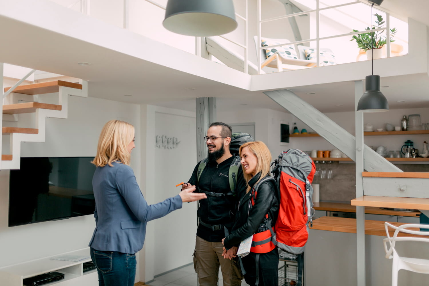 Une femme vêtue d'un blazer bleu et d'un jean remet une clé à un couple souriant, sacs à dos sur les épaules, à l'intérieur d'une maison moderne.
