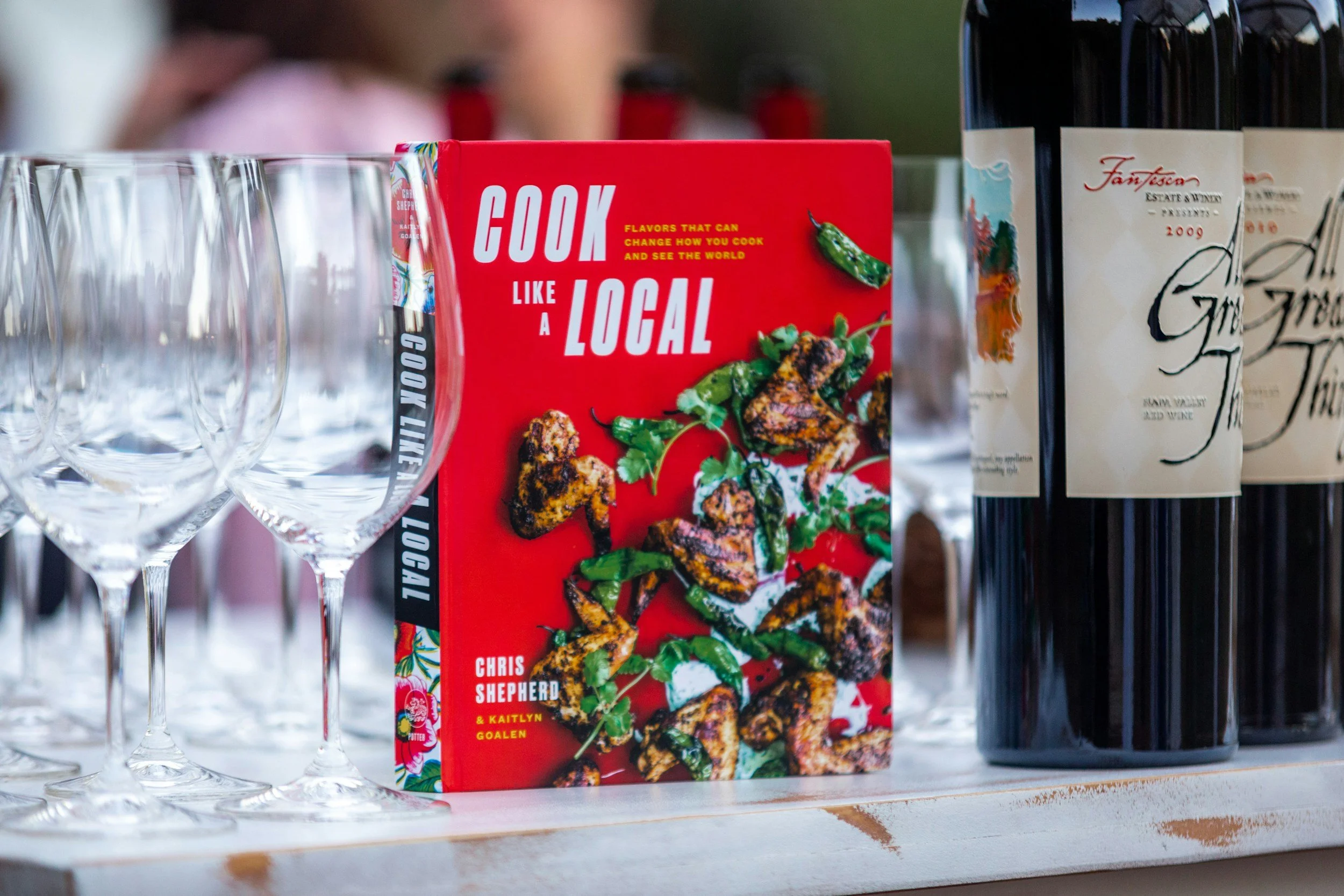 A cookbook titled 'Cook Like a Local' by Chris Shepherd and Kaitlyn Goalen stands among empty wine glasses and bottles of wine on a table, with a blurred background.
