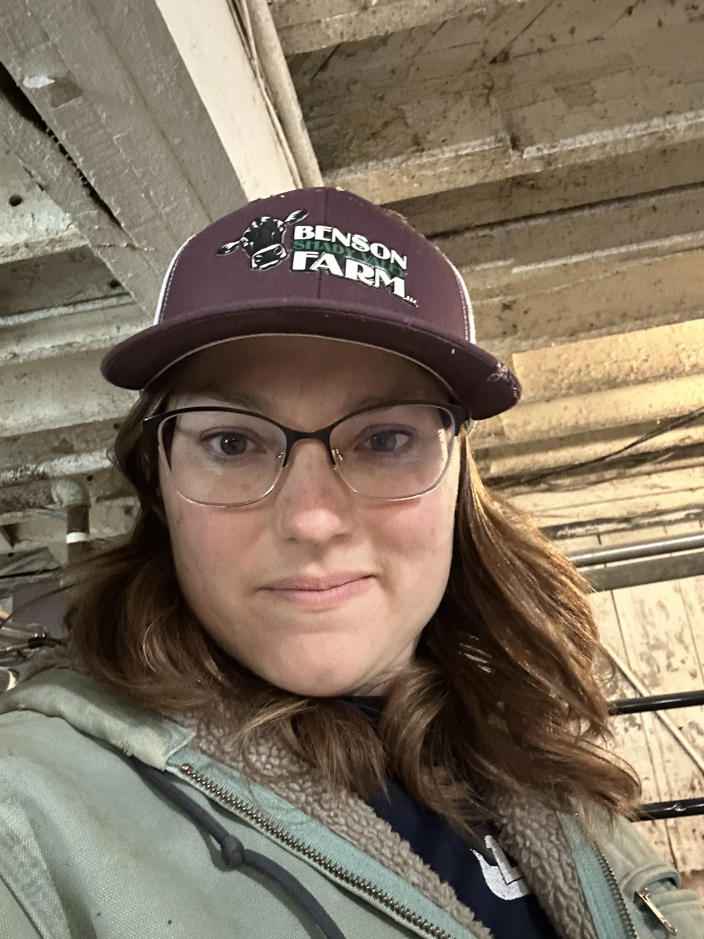 A woman wearing glasses, a maroon Benson farm cap, and a green jacket standing in a rustic, unfinished building