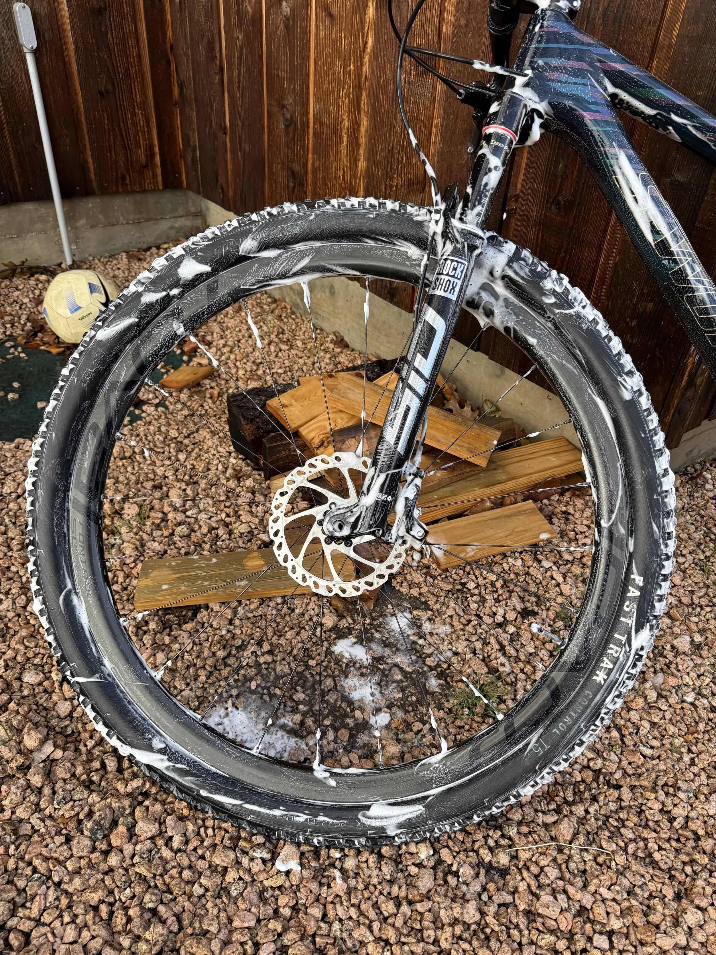 A mountain bike wheel with soapy foam on the tire, rim, and spokes, leaning against a wooden fence, with a soccer ball on the gravel ground nearby.