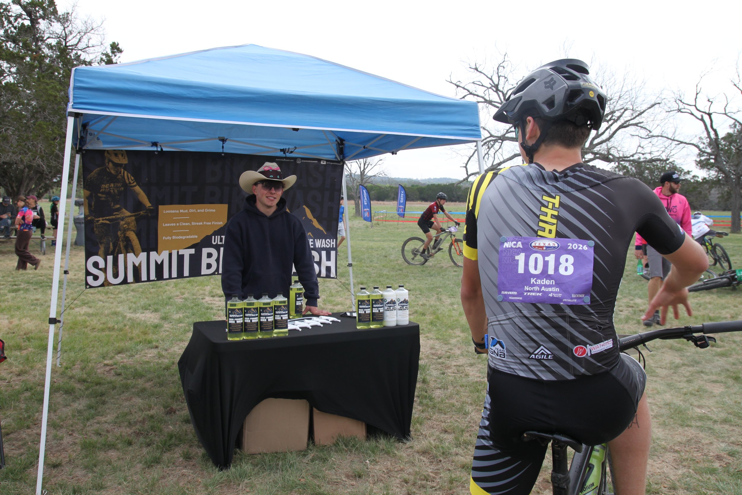 A cyclist with a race number 1018 stands with his back to the camera, talking to a person at a booth with bike wash products at an outdoor event. The booth has a blue canopy, and there are other cyclists and spectators in the background.