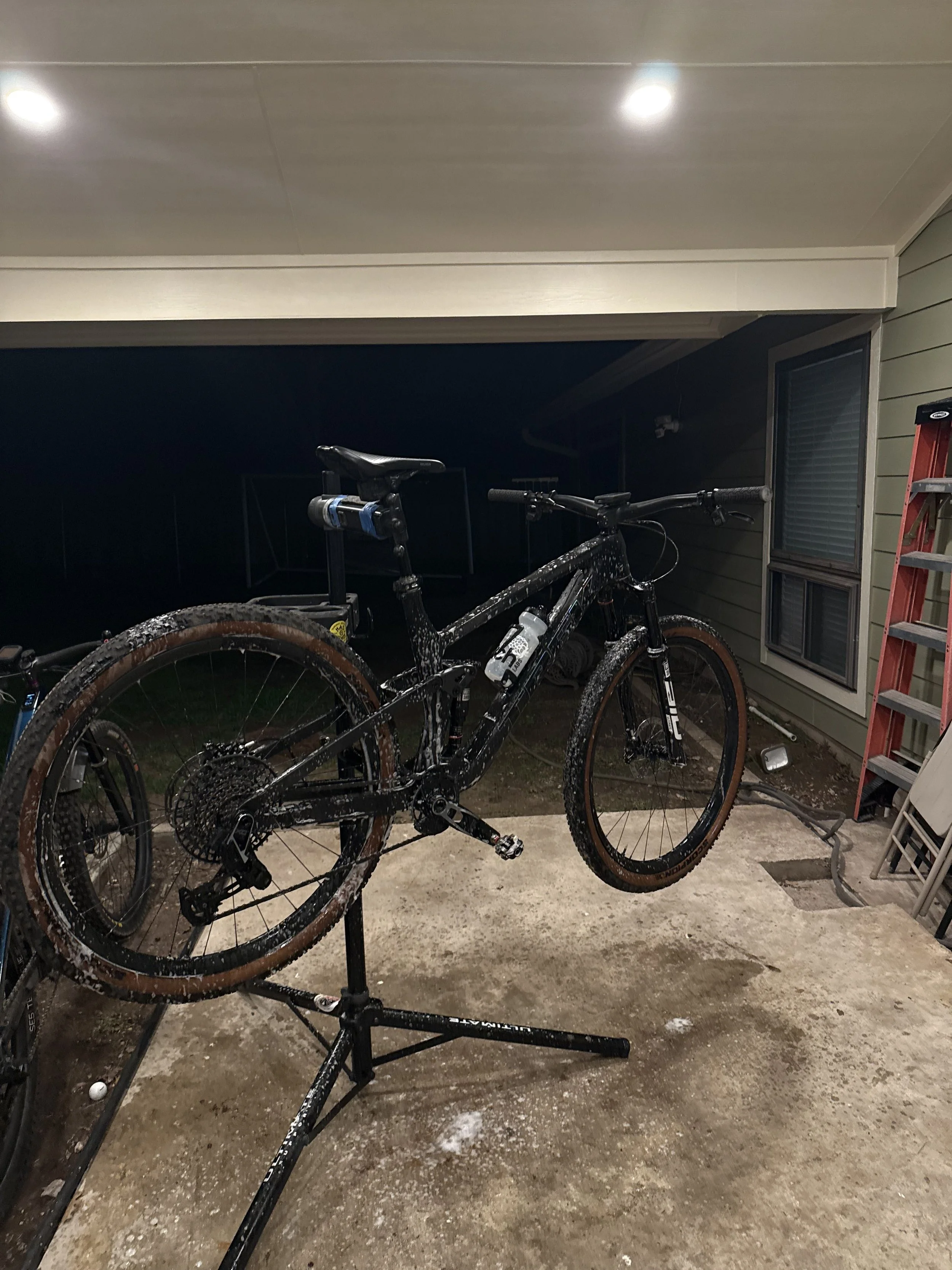 A black mountain bike with mud on the tires and frame, placed on a bike stand inside a garage or basement, with a red ladder nearby and fluorescent ceiling lights above.