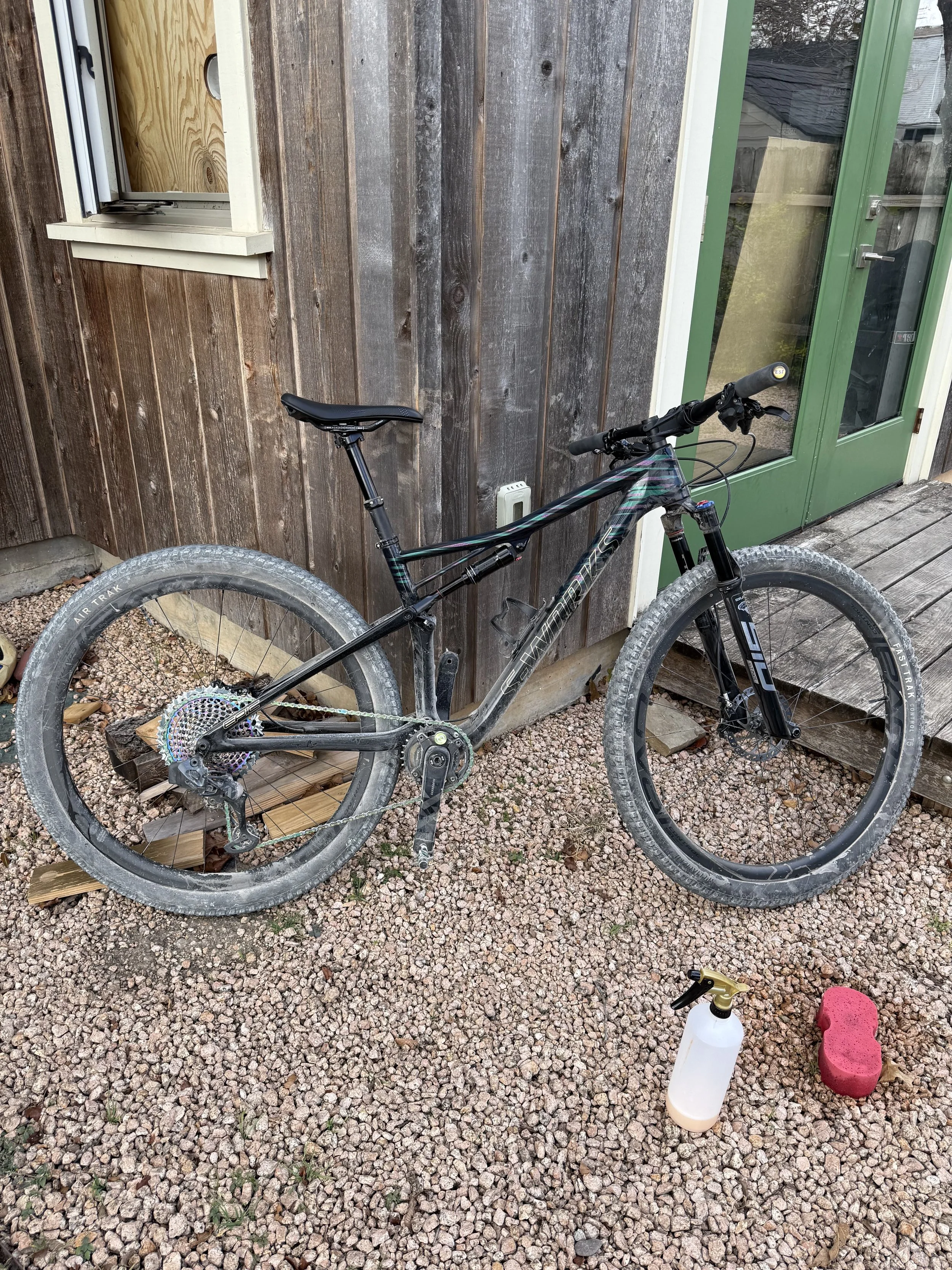 A black mountain bike leaning against a rustic wooden wall outside a house, with cleaning supplies including a spray bottle, a pink sponge, and a spray nozzle on the gravel ground nearby.