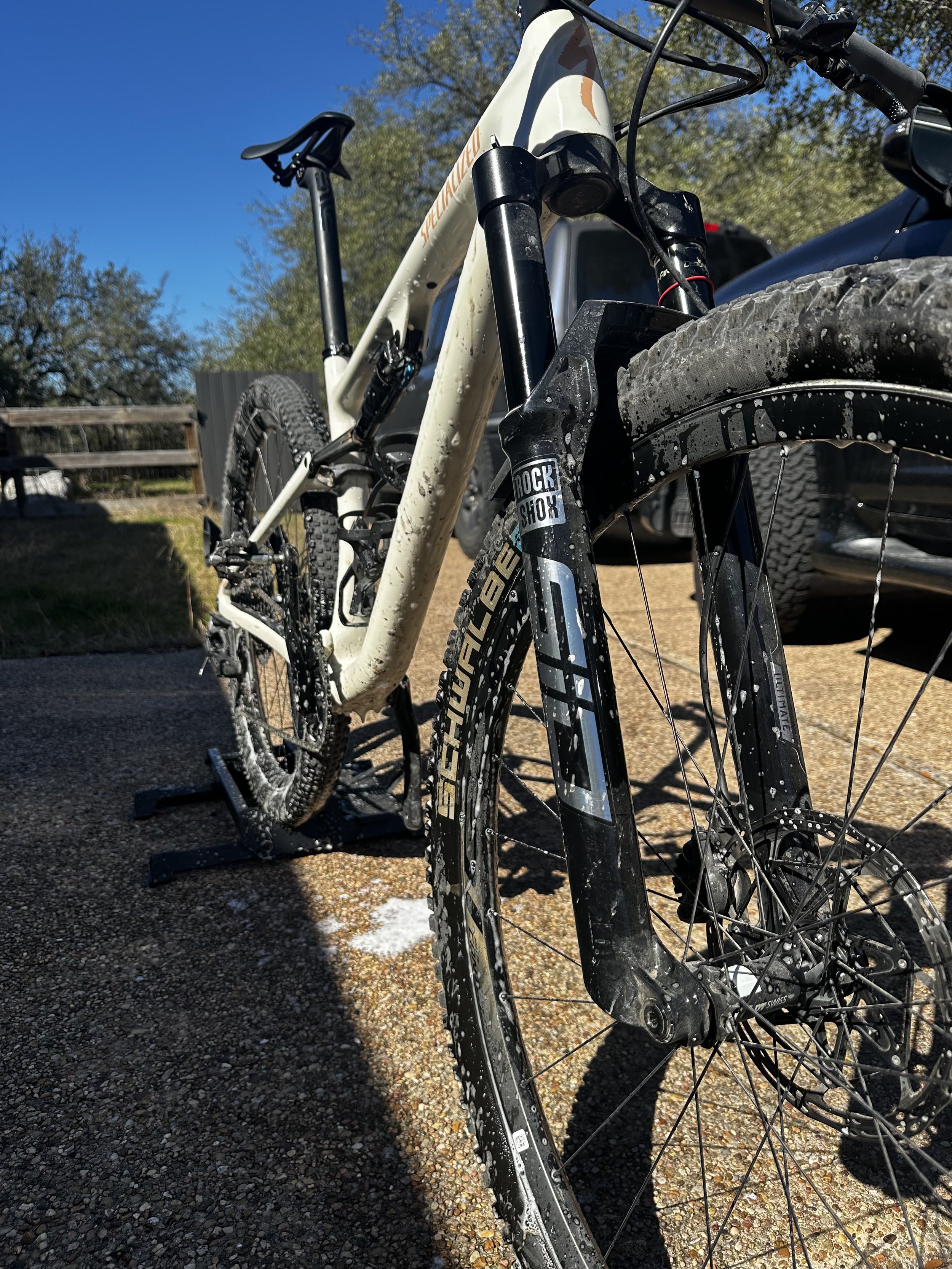 Close-up of a mountain bike with mud-splattered tires and suspension, parked on a gravel surface on a sunny day with trees and a vehicle in the background.