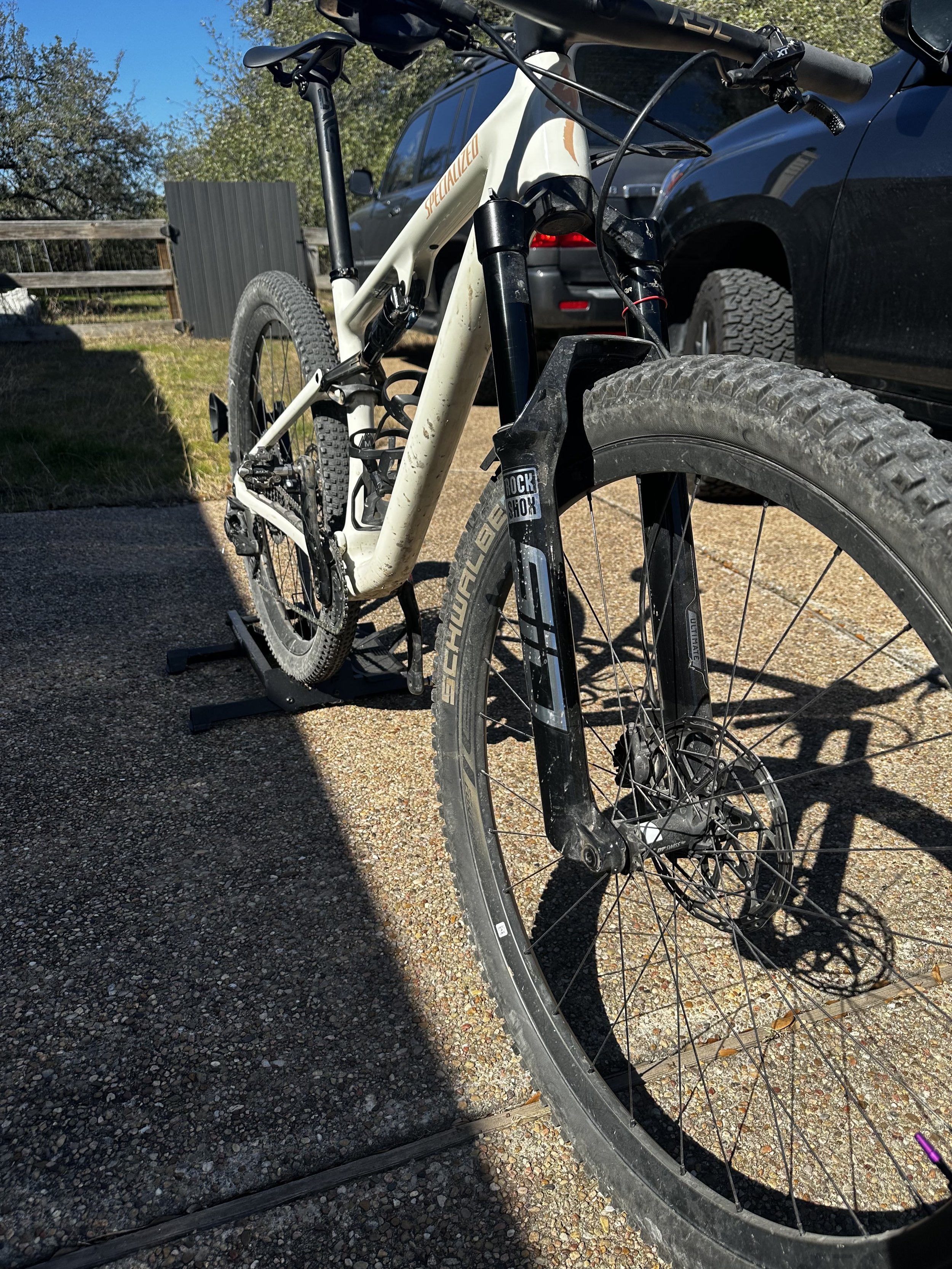 A close-up of a mountain bike with a white frame and brand label, parked on a rough concrete surface next to a black vehicle. The bike features a front suspension fork, knobby tires, and gear components. Sunlight and shadows are visible.