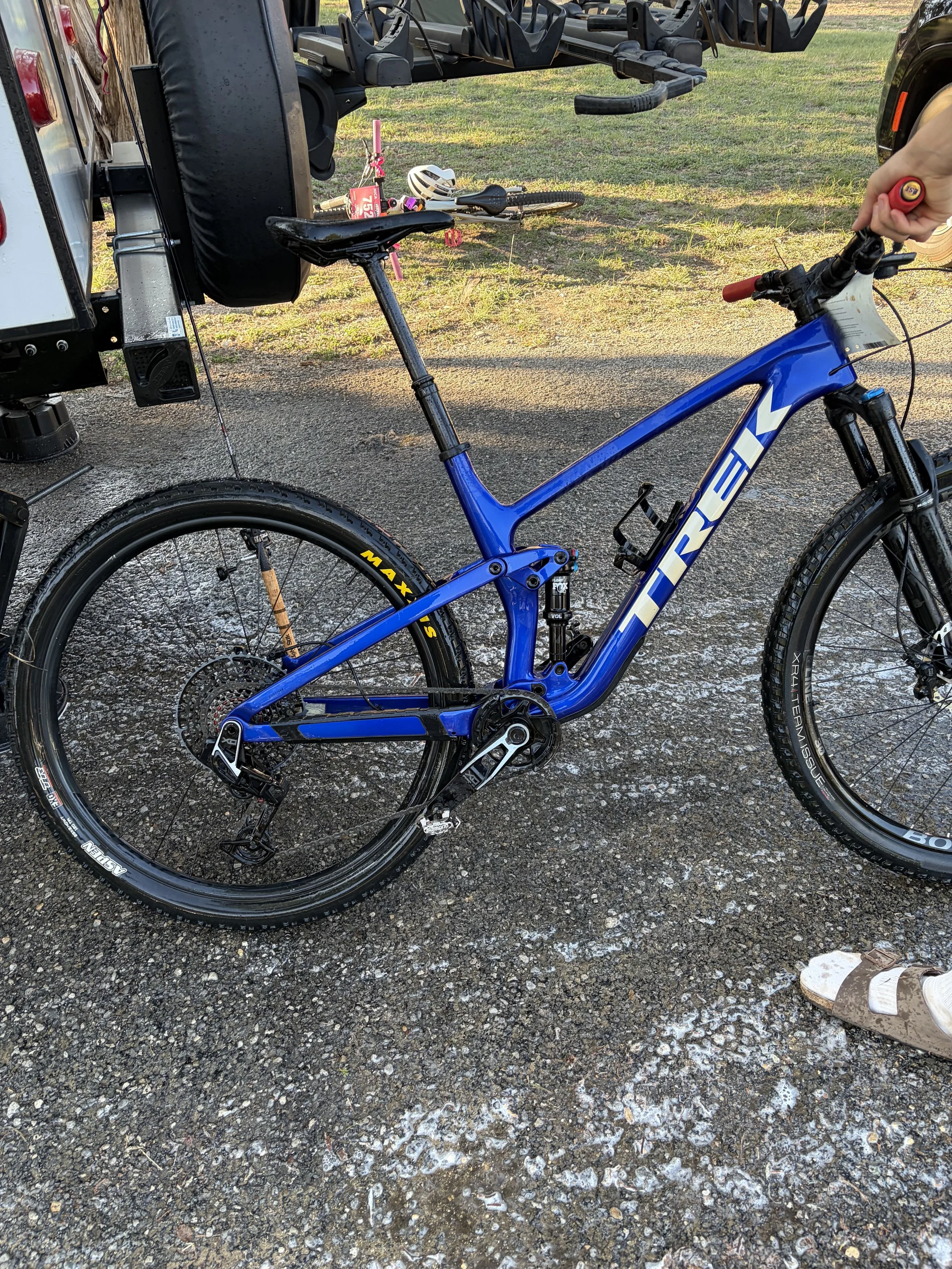 A blue Trek mountain bike with disc brakes and a rear suspension, parked on a wet, gravelly surface. A person's hand is holding a red tool near the bike's handlebar. In the background, visible are a bicycle helmet, a pink children's bike with trainin