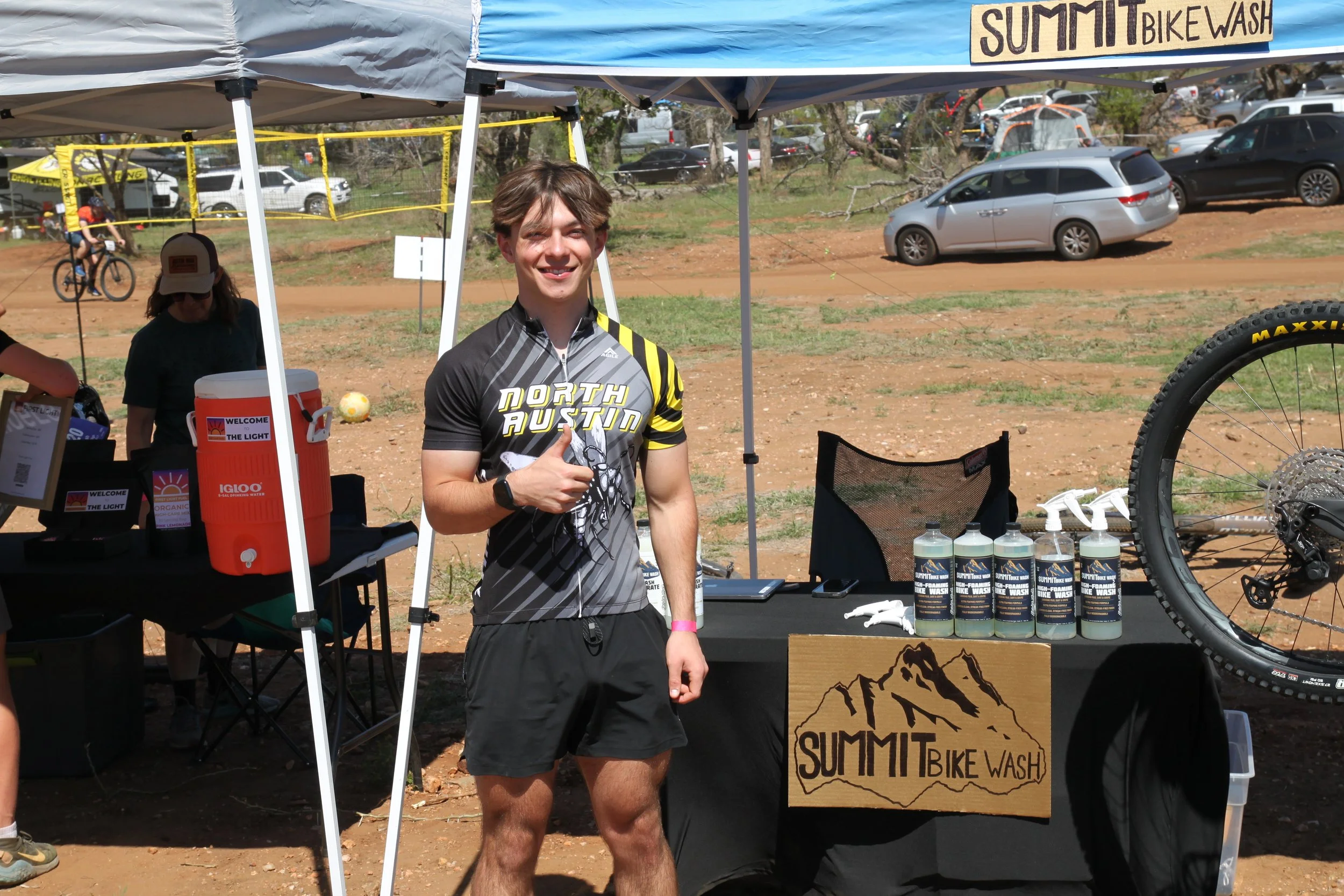 A young man in cycling gear standing at a mountain bike wash station, giving a thumbs up. The station has bottles of bike wash, a sign that reads 'Summit Bike Wash,' and is set outdoors with parked cars and a dirt trail in the background.