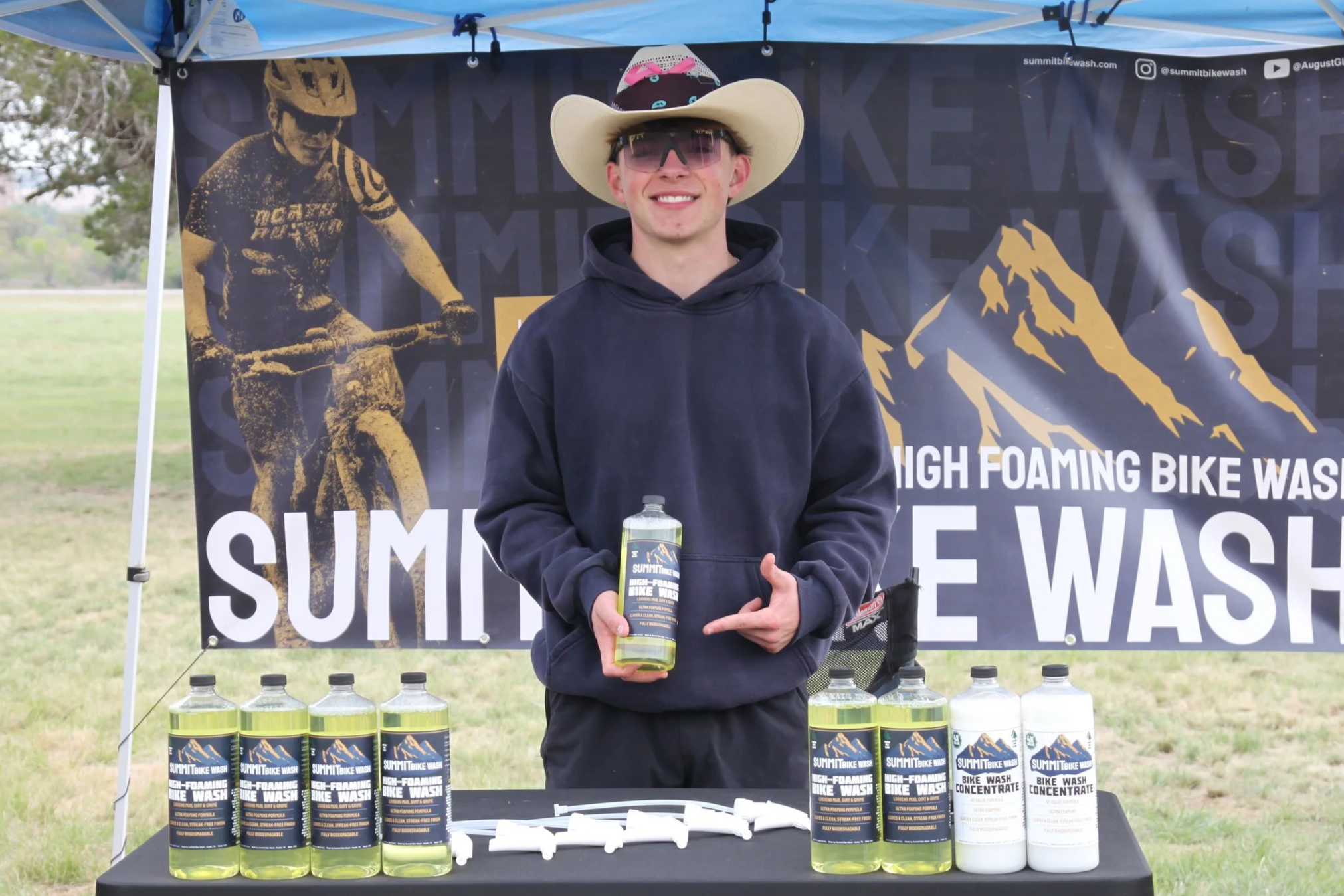 A young man wearing a cowboy hat, sunglasses, and a dark hoodie holding a bottle of bike wash at an outdoor booth. The booth features a banner and several bottles of bike cleaning product labeled 'Summit Bike Wash' in front of him.