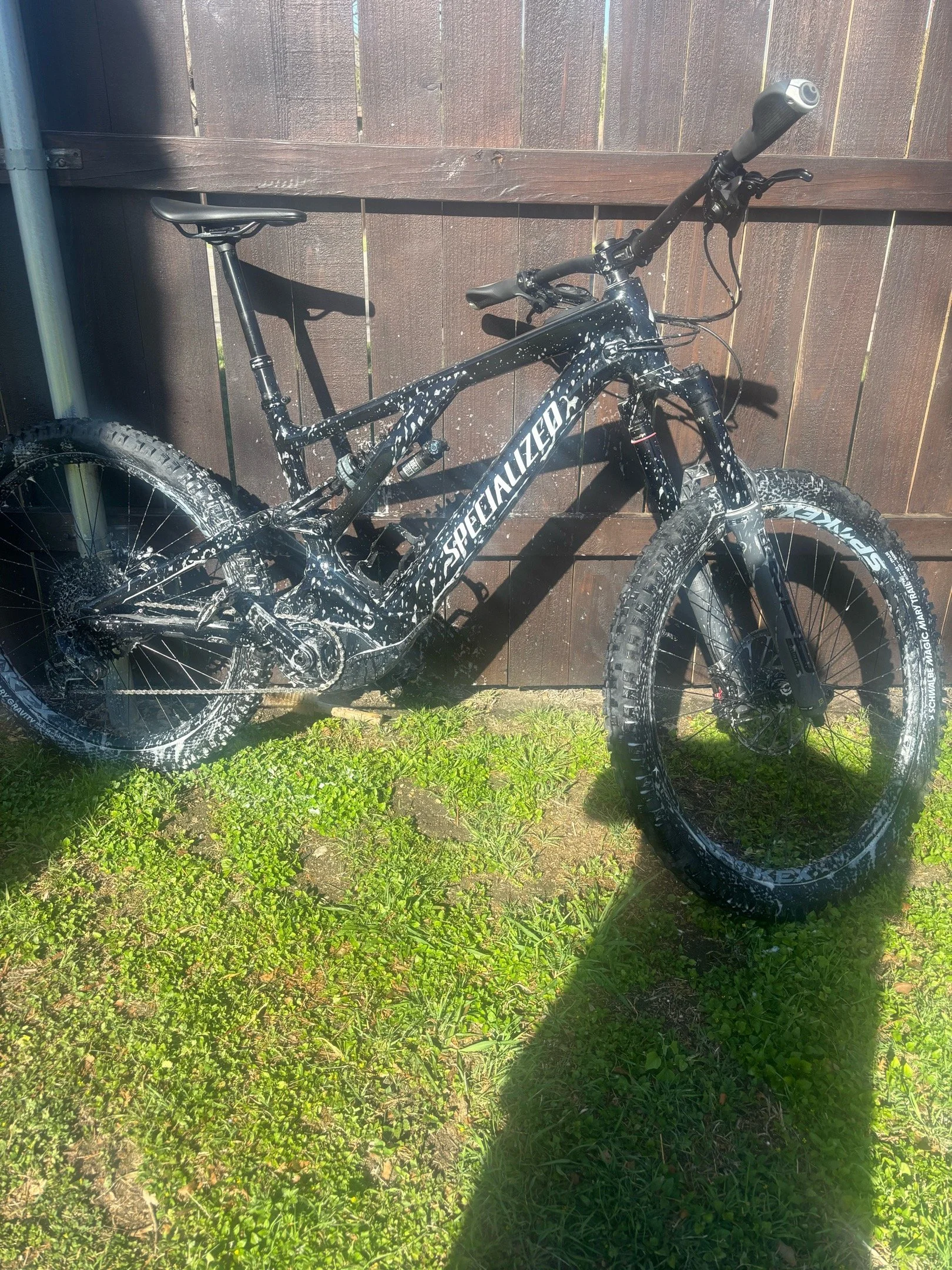 Black mountain bike with speckled white paint, leaning against a wooden fence on grass and dirt ground, with sunlight casting shadows.