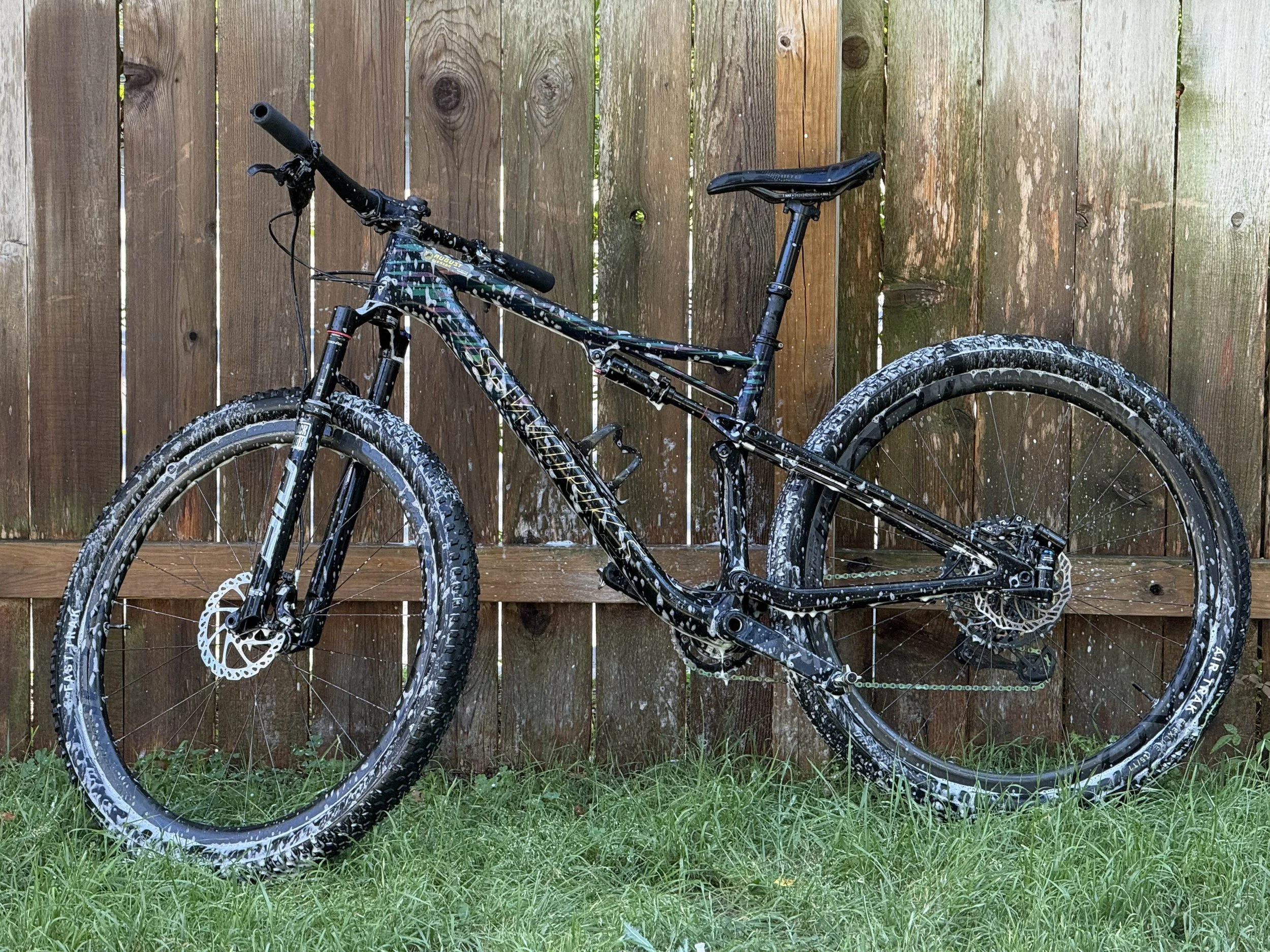 A mountain bike with black frame and colorful patterns, leaning against a wooden fence, with muddy tires and grass in the foreground.