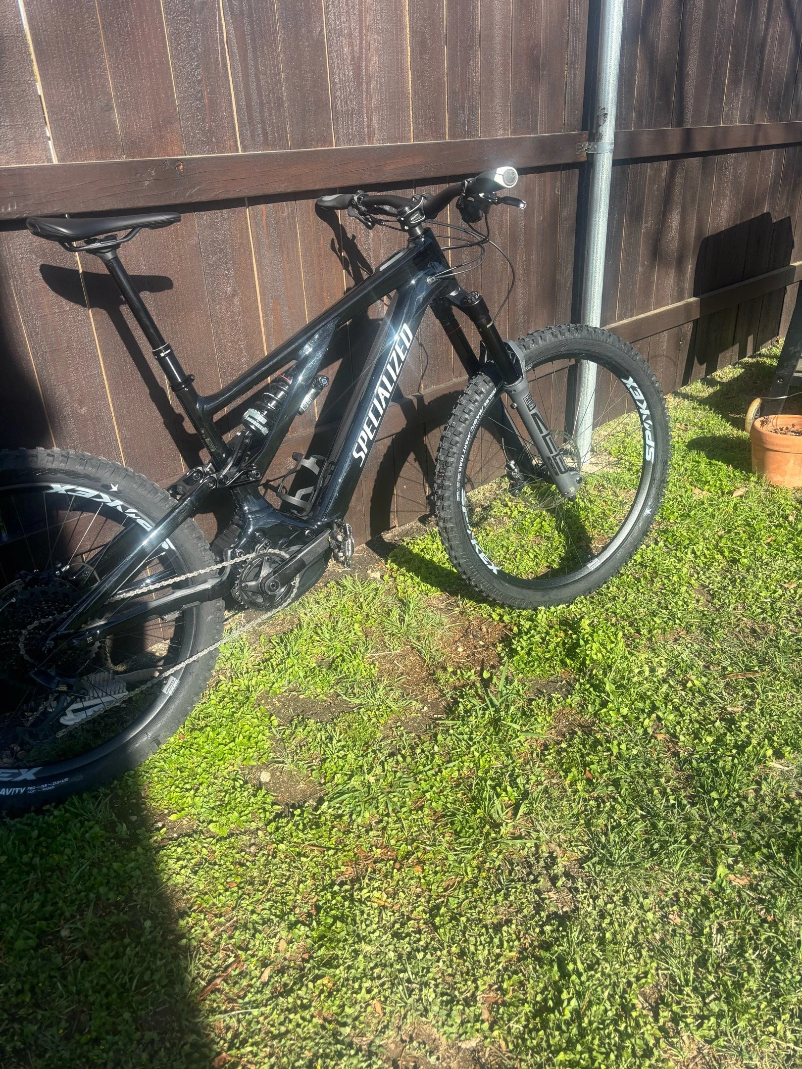 A black Specialized mountain bike is leaning against a wooden fence with vertical planks, sunlight casting shadows, and a small pot of plants on the ground near the fence, with grass and dirt around.