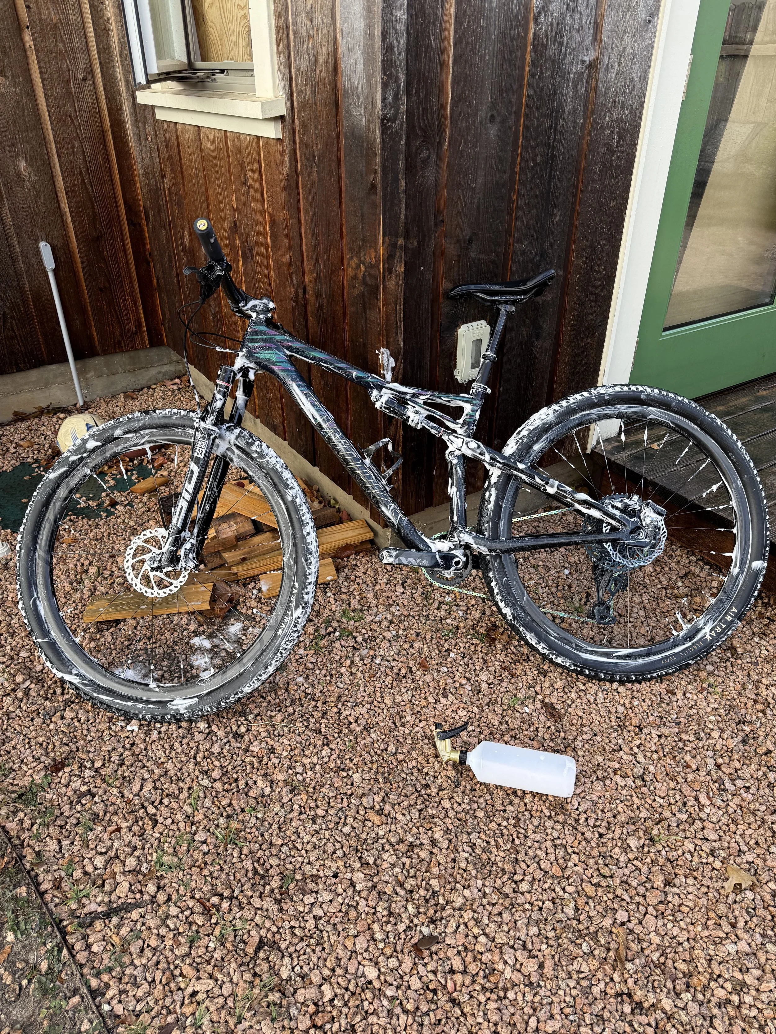 A mountain bike with white soap suds on it leaning against a dark brown wooden wall outside a house. The ground is covered with small reddish-brown gravel and there are objects such as a white spray bottle, a small white object, and a green door visi