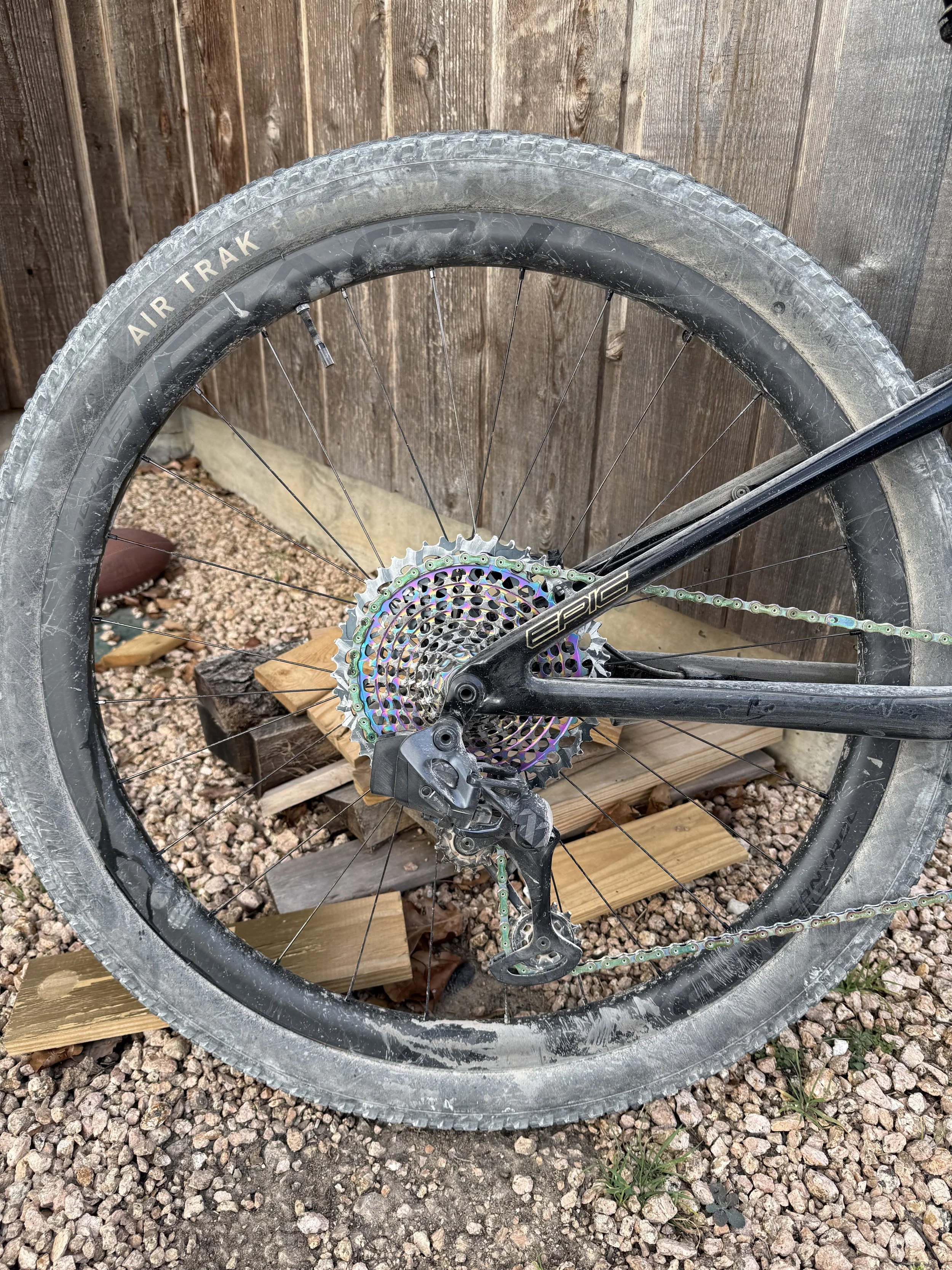 A mountain bike tire and rear derailleur are shown next to a wooden fence and gravel ground, with small plants growing nearby.