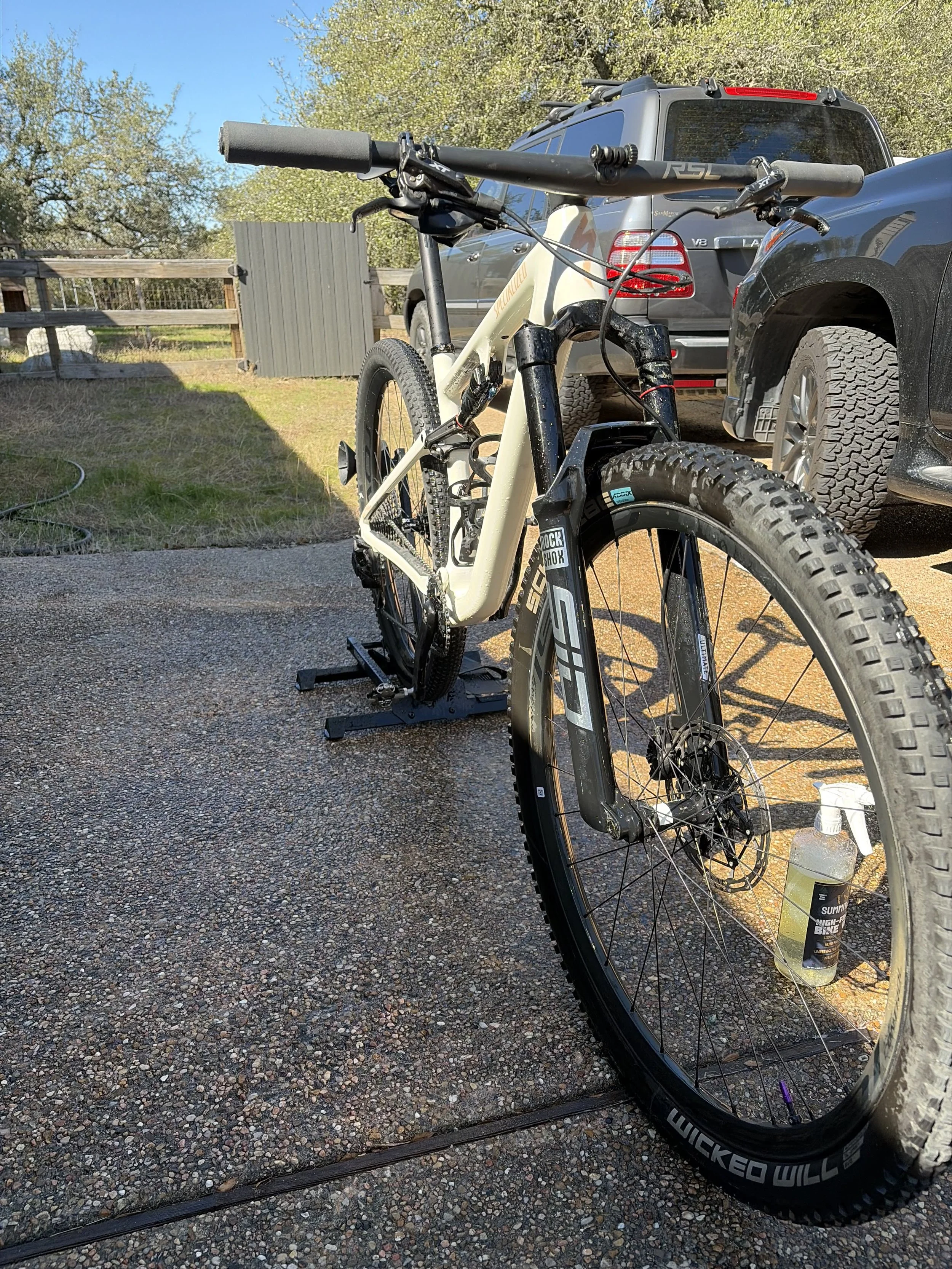 A mountain bike mounted on a trainer outdoors with a building and parked cars in the background.
