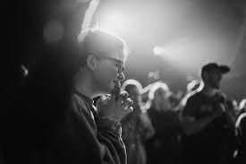 A person praying or reflecting with their hands clasped in a dark, crowded setting, illuminated by soft light.