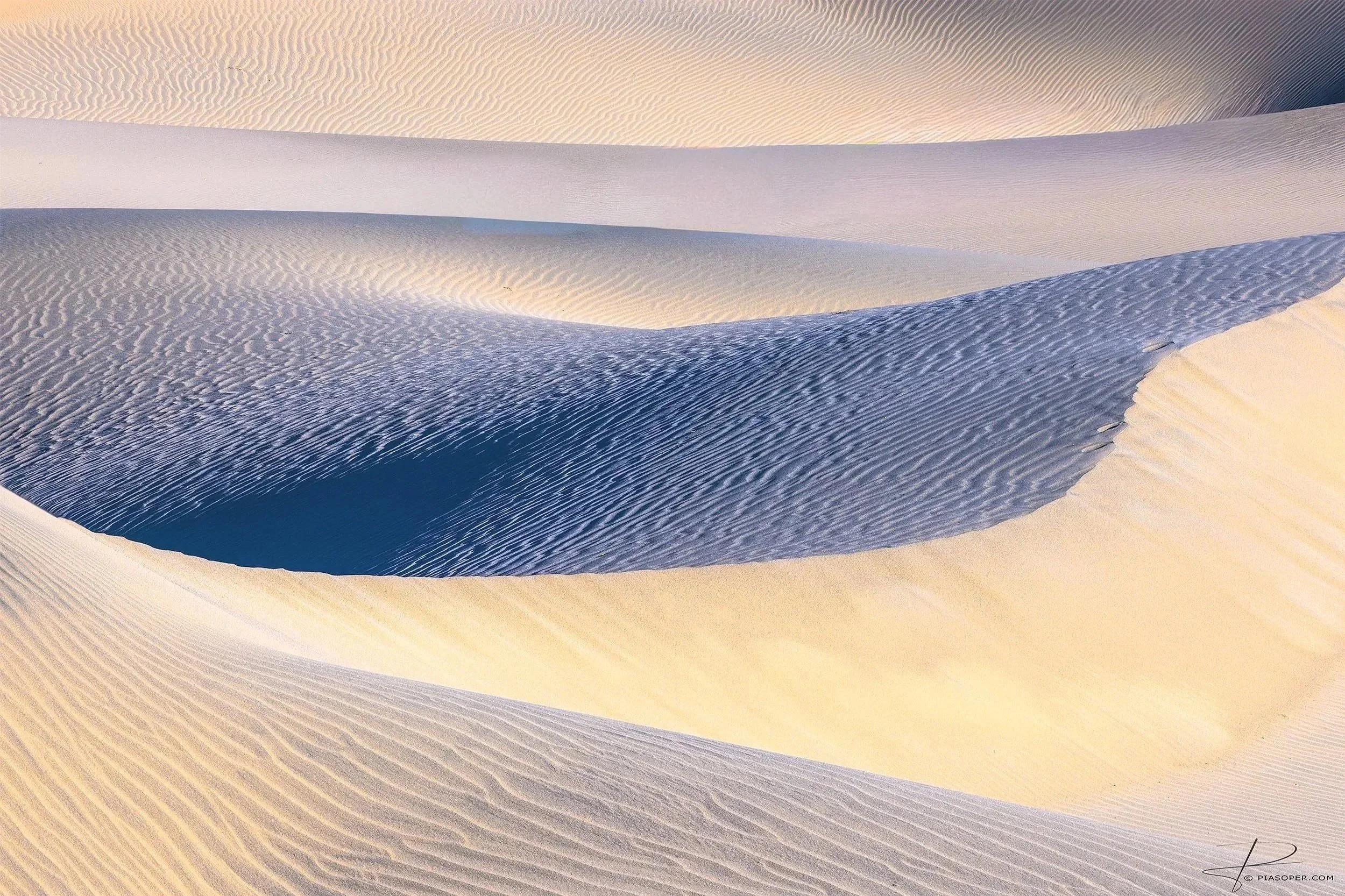 Soft morning light ascends over dunes and ripples in pastel shades of blue, yellow, magenta tones. Valleys descend into darker tones and the dunes dance like waves creating shadows of light and dark at Mesquite Dunes in Death Valley National Park