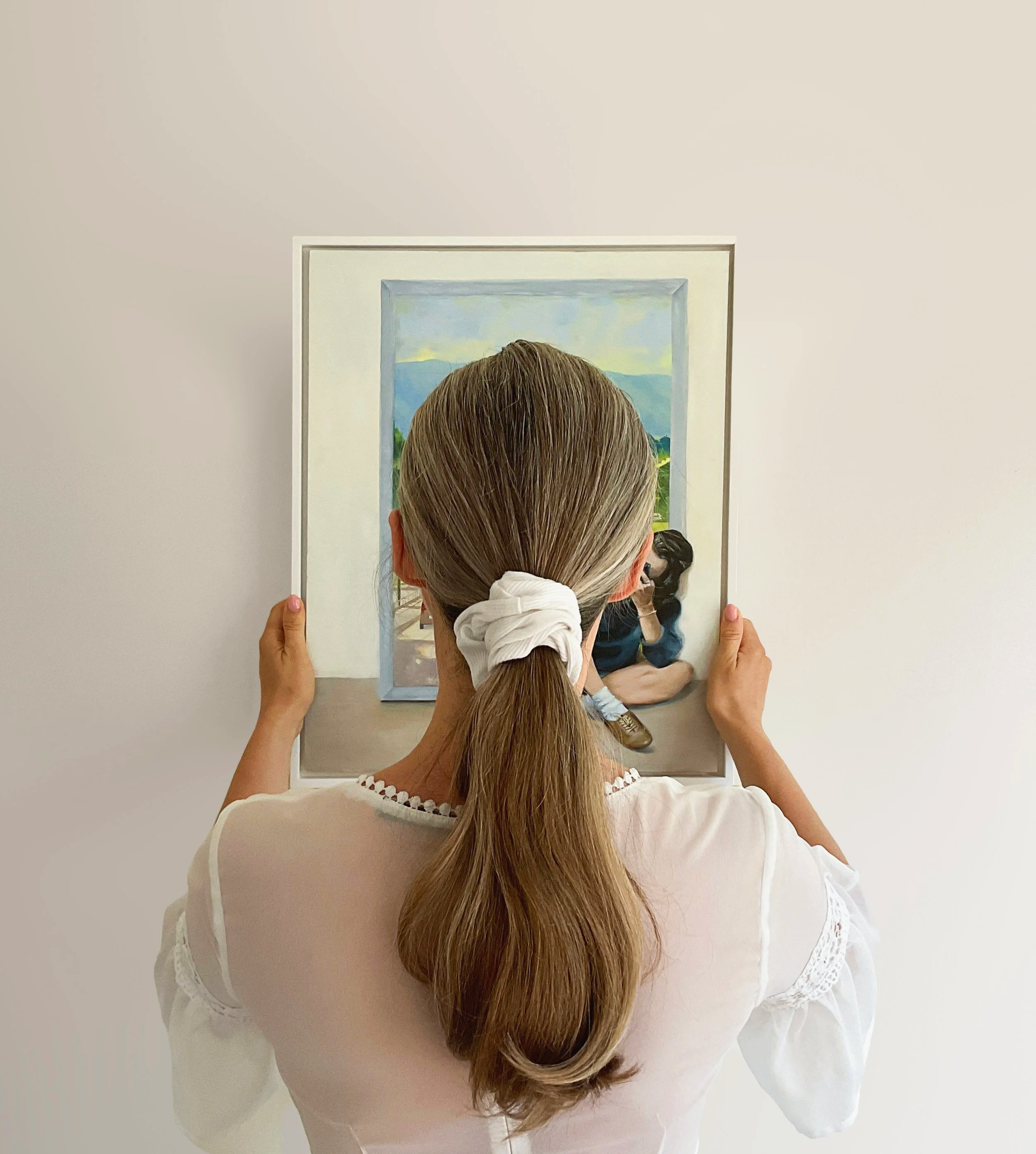 A woman with long, brown hair tied with a white scrunchie, seen from behind, holding a mirror and looking at her reflection, with a plain light-colored wall in the background.