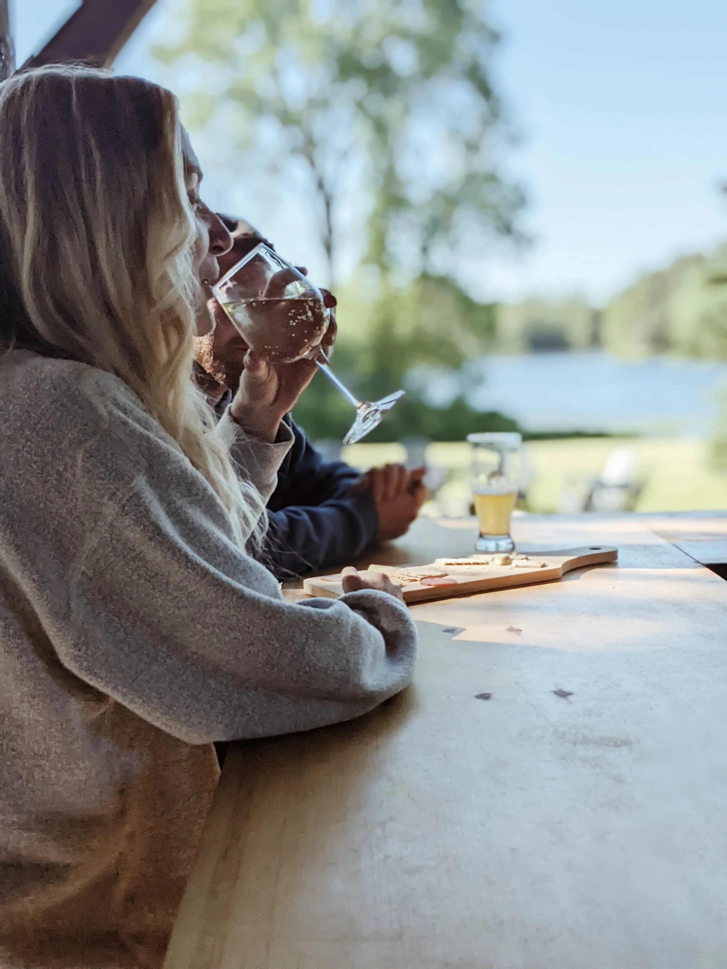 Three people sitting at a wooden table outdoors by a lake, drinking wine and beer, with a board and glasses, with nature and trees in the background.
