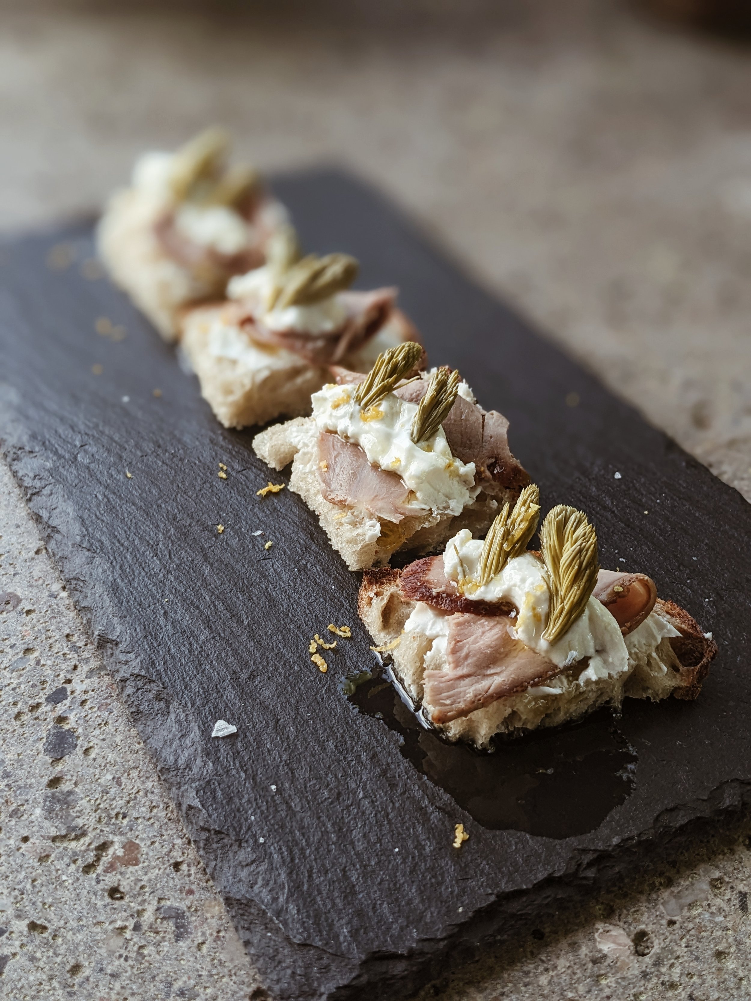 Close-up of canapé with bread, sliced meat, dollop of cream, and artichoke garnishes on a dark stone serving board.
