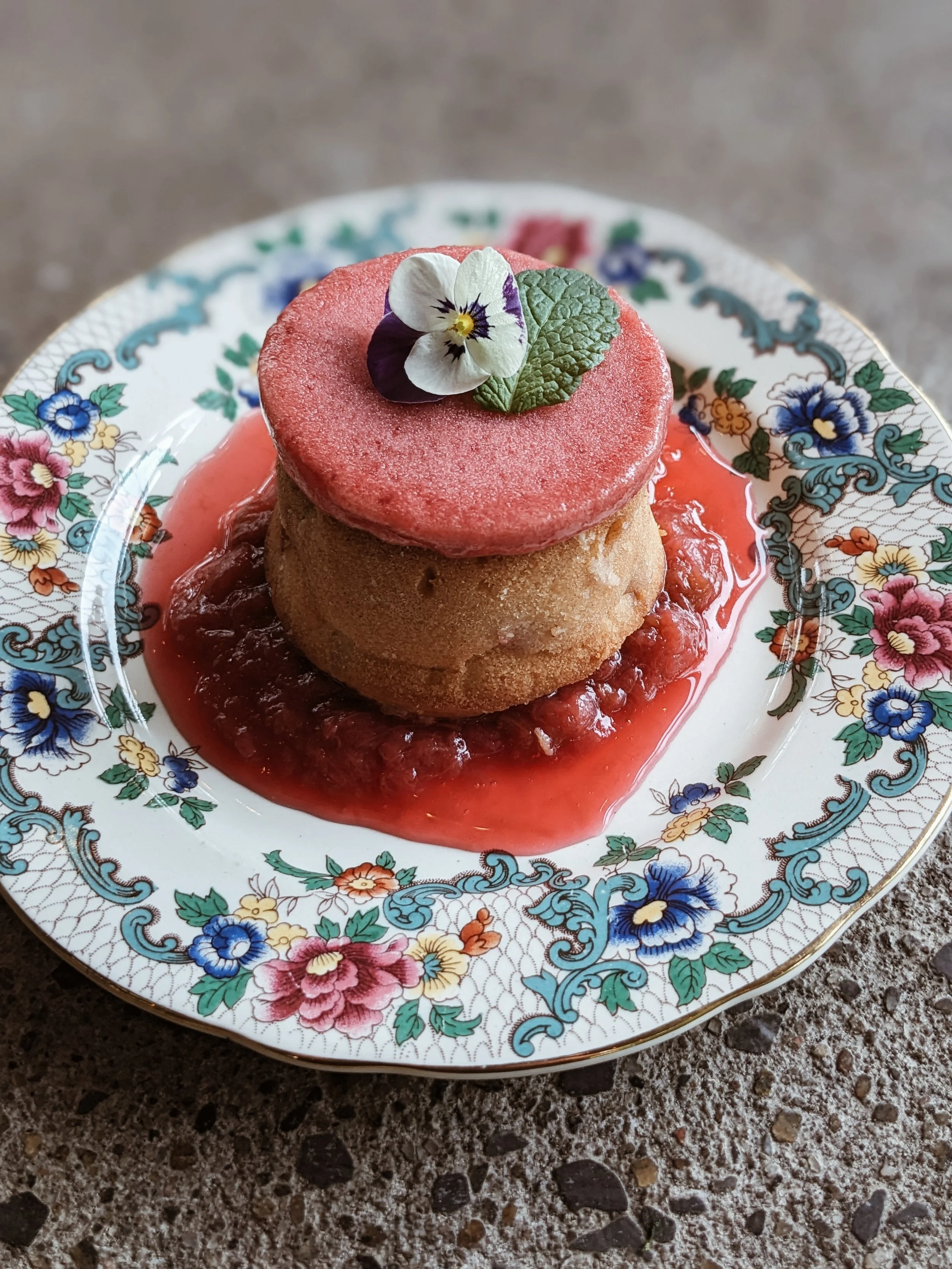 Dessert with a round baked good topped with a pink flower, green mint leaf, and a pink icing disc, served on a floral-patterned plate with fruit compote underneath.
