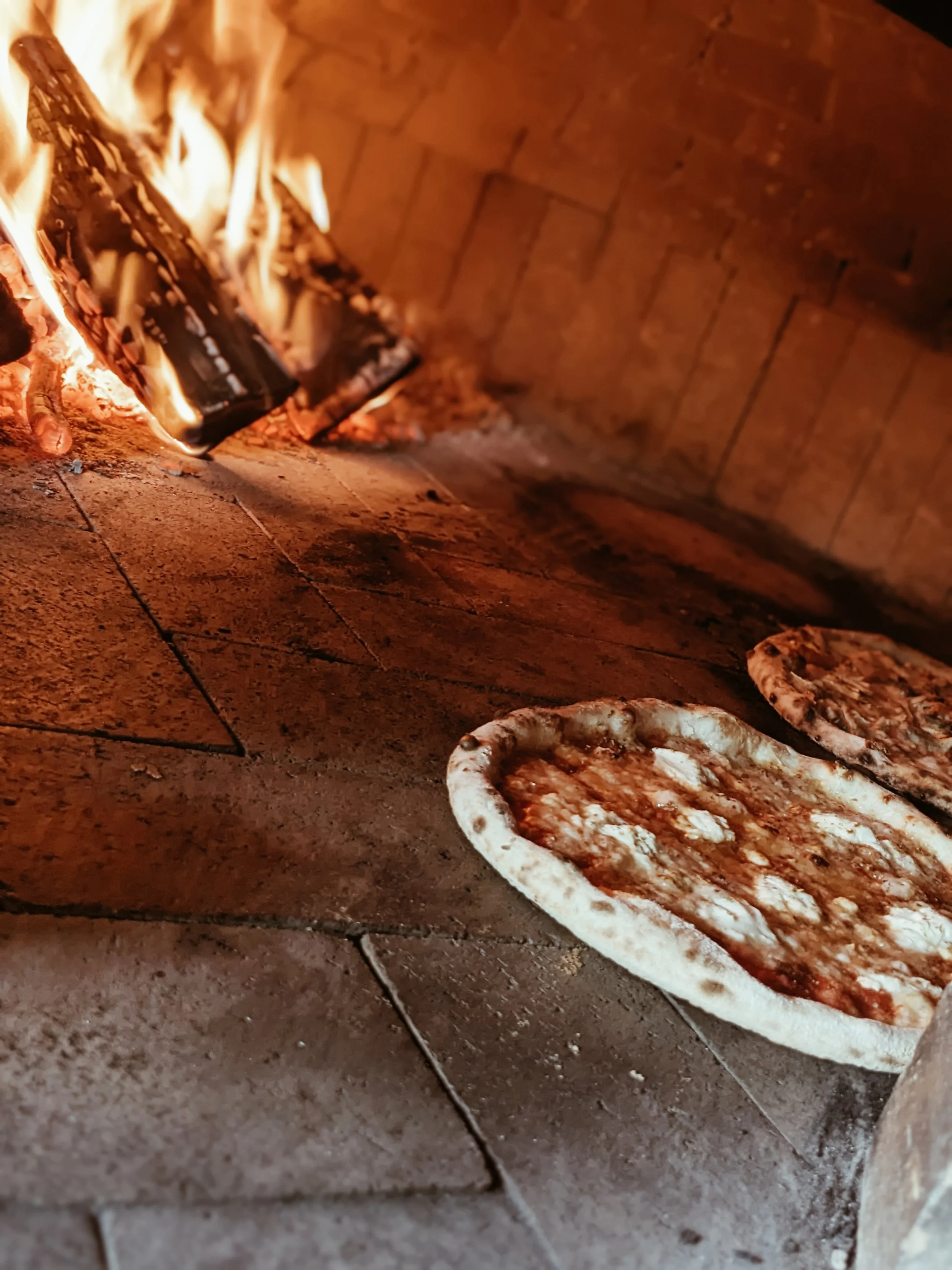 Pizza baking in a wood-fired oven with flames visible in the background.