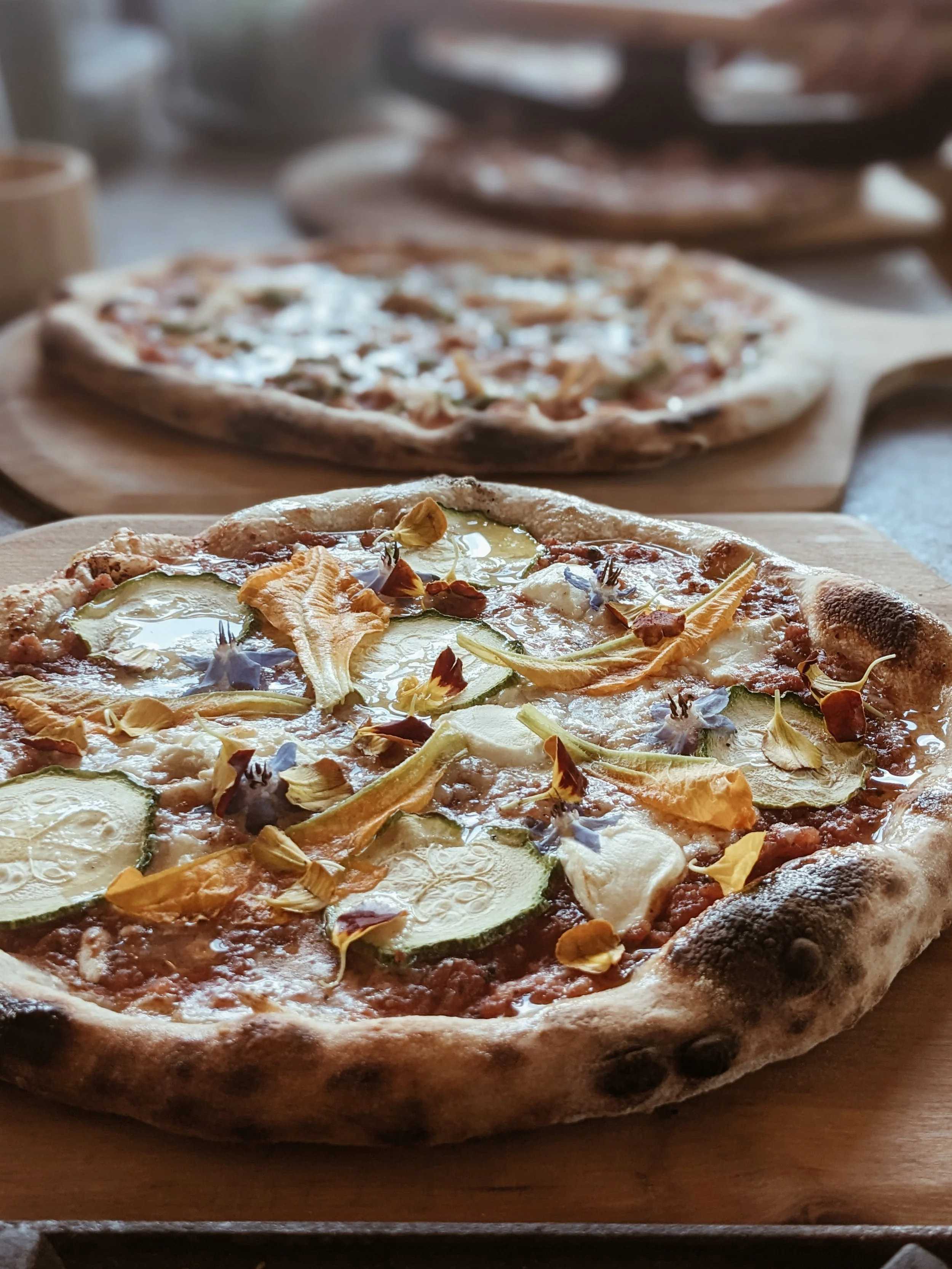 Close-up of a freshly baked pizza topped with zucchini slices, cheese, and edible flower petals on a wooden serving board. Two other pizzas are visible in the background.