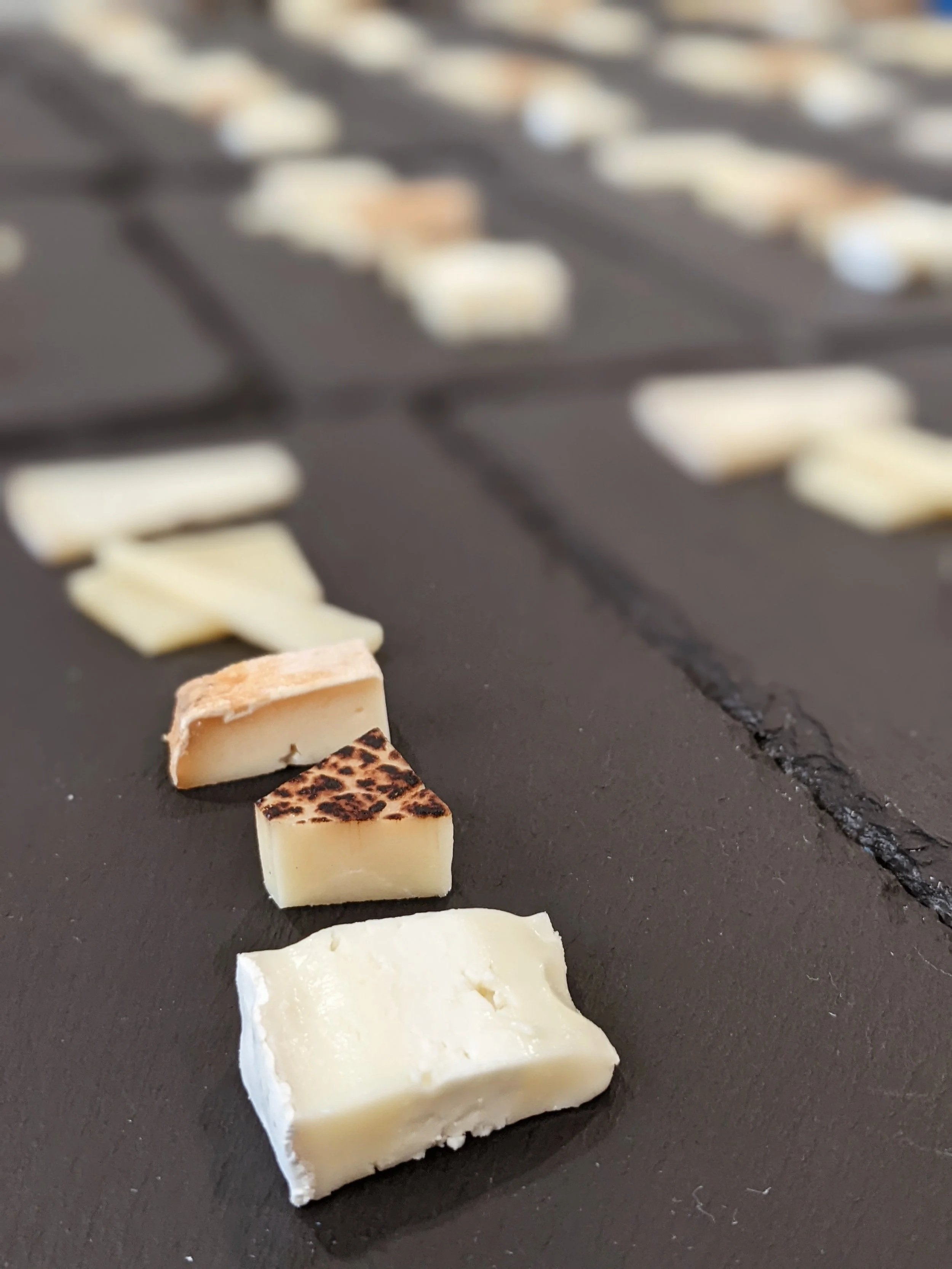 Close-up of three pieces of cheese in front of a cheese board with sliced cheese in the background.