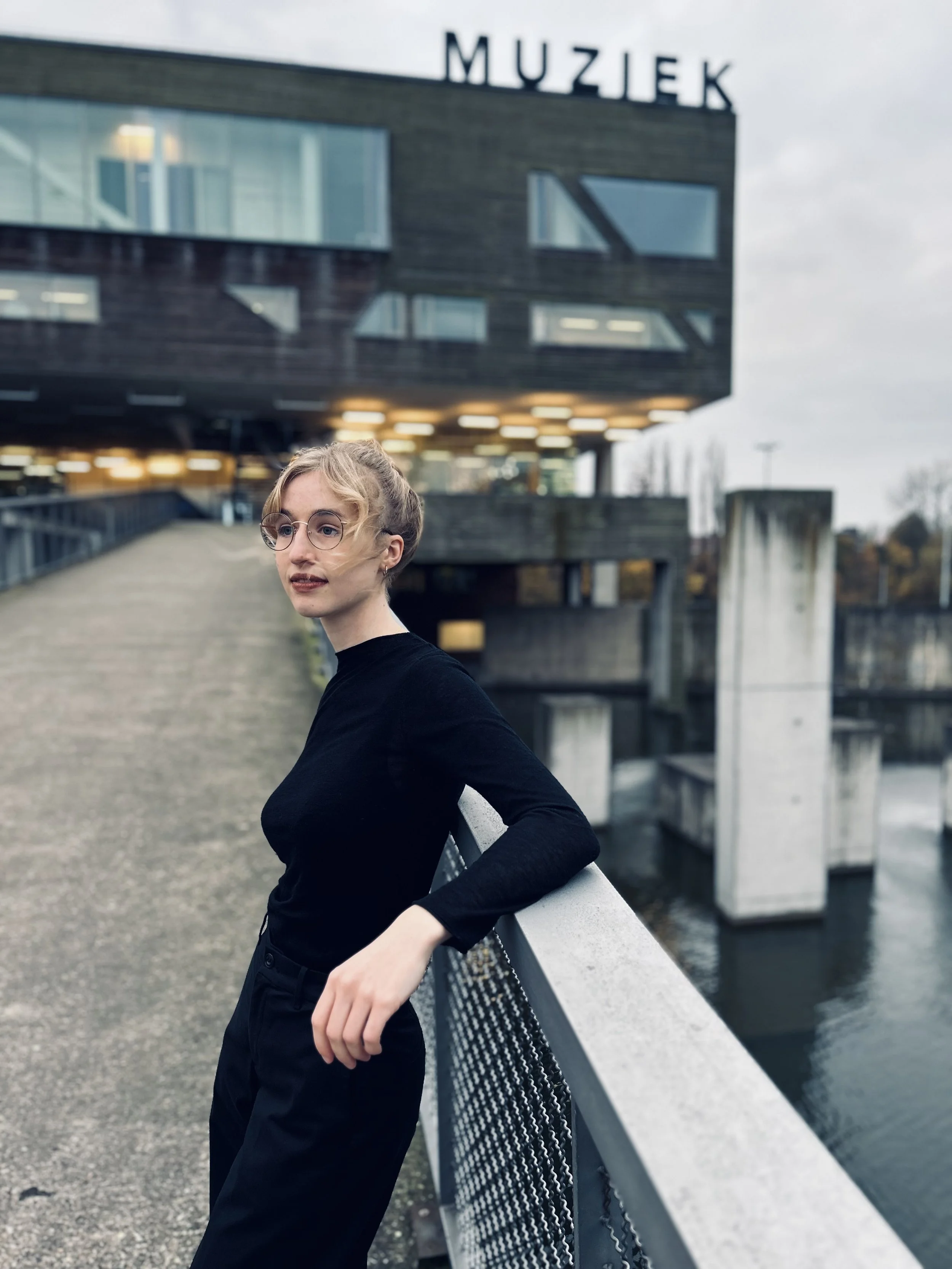 A woman with short blonde hair, wearing glasses and a black outfit, leaning on a railing on a bridge near a modern building with the word 'MUZIEK' on it, over a body of water on a cloudy day.