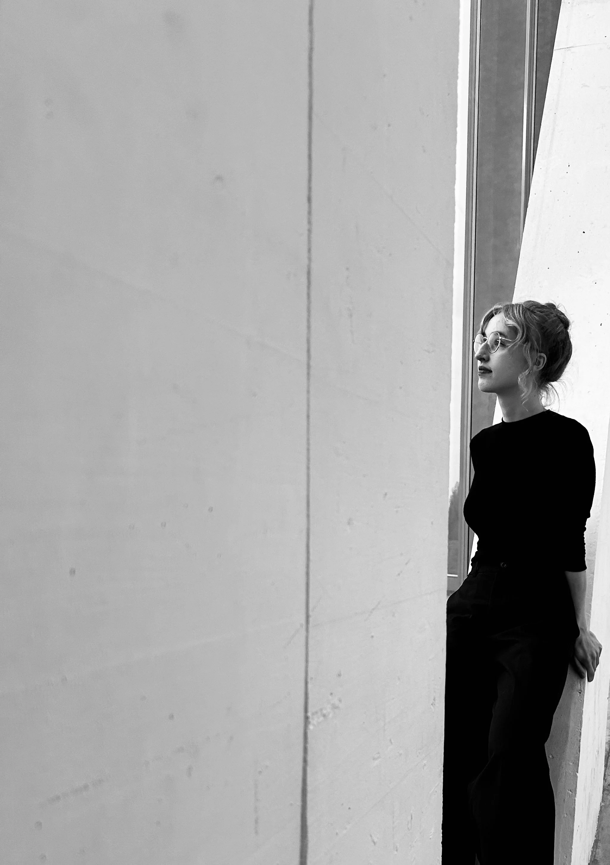 Black and white photo of a woman with glasses, dark hair in a bun, dressed in black, leaning against a white wall near a window, looking to the right.