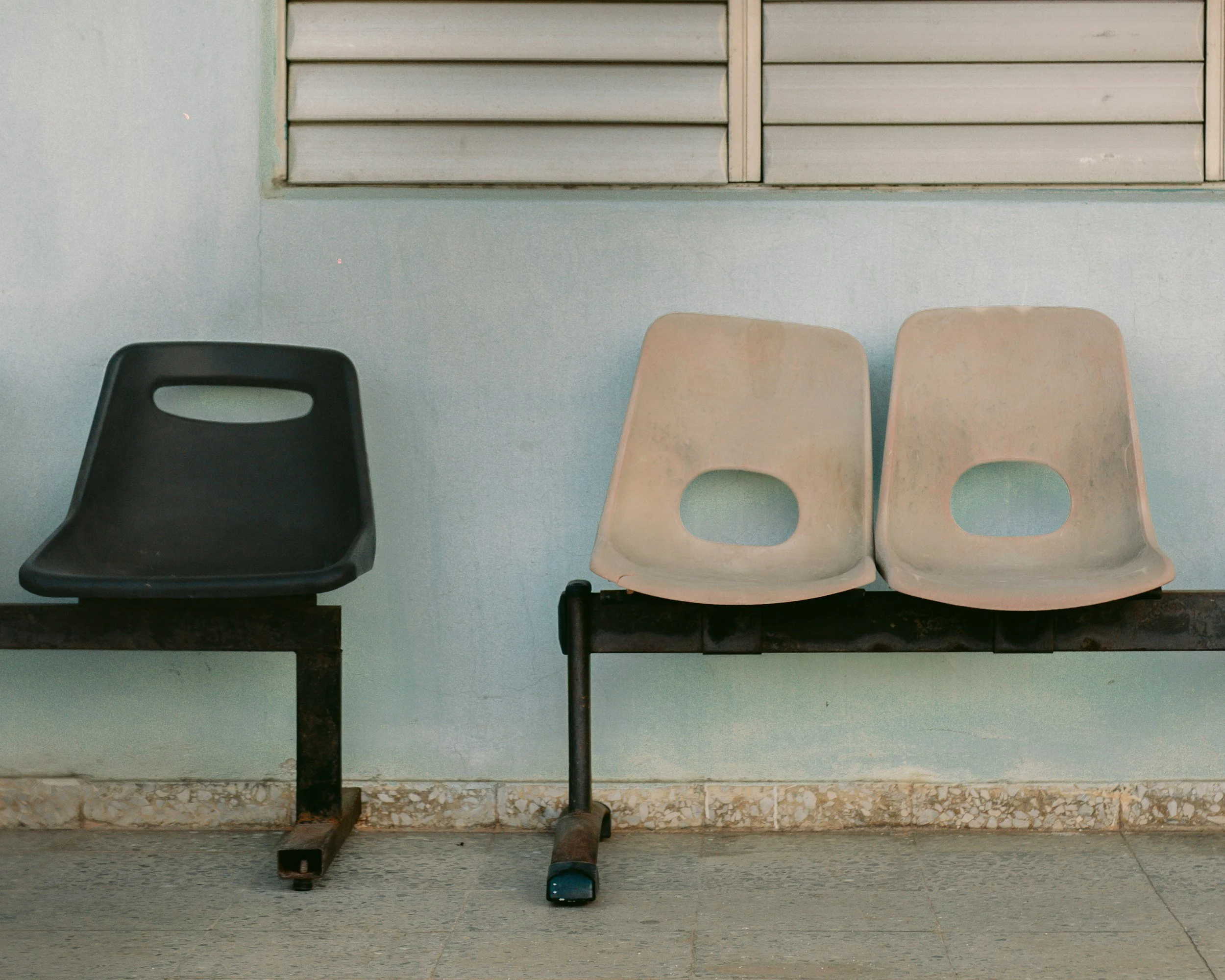 Three old and worn chairs, black and cream-colored, sitting on a concrete floor against a pale blue wall with a window.