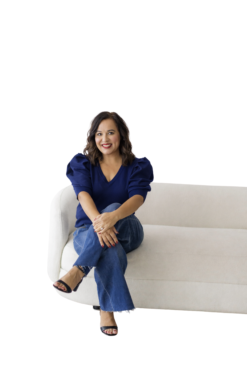 A woman sitting on a white couch, smiling, wearing a blue blouse and blue jeans, with dark hair and red lipstick.