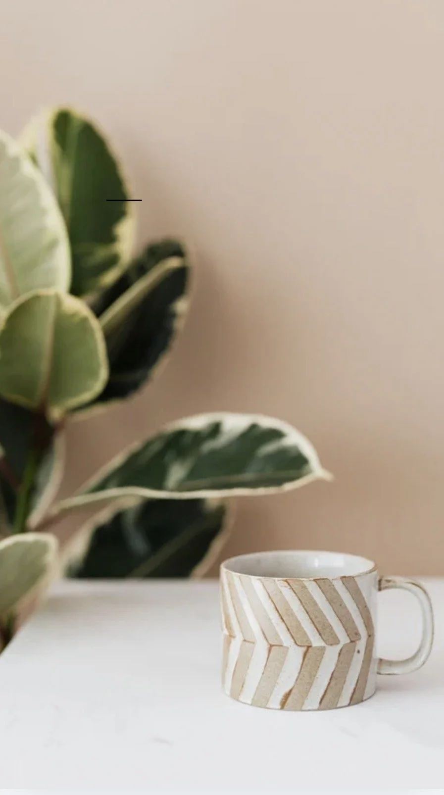 A white mug with beige diagonal stripe pattern placed on a white surface with a large green leafy plant in the background.