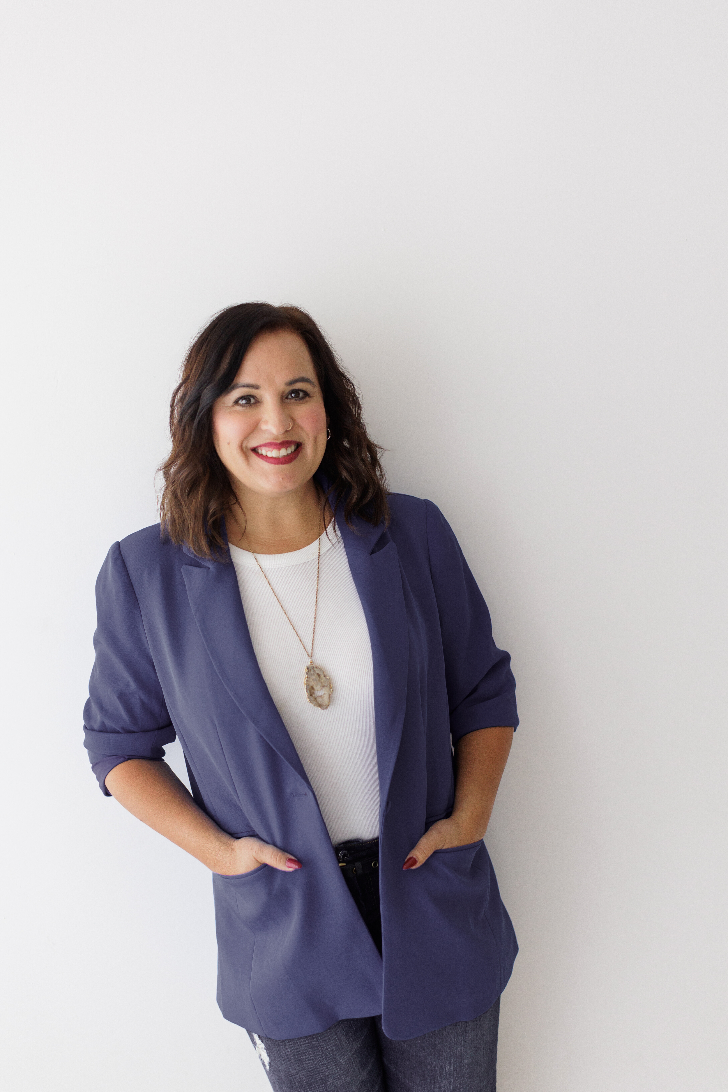 A woman with shoulder-length dark hair, wearing a navy blazer over a white top, smiling and standing with her hands in her pockets against a plain white background.