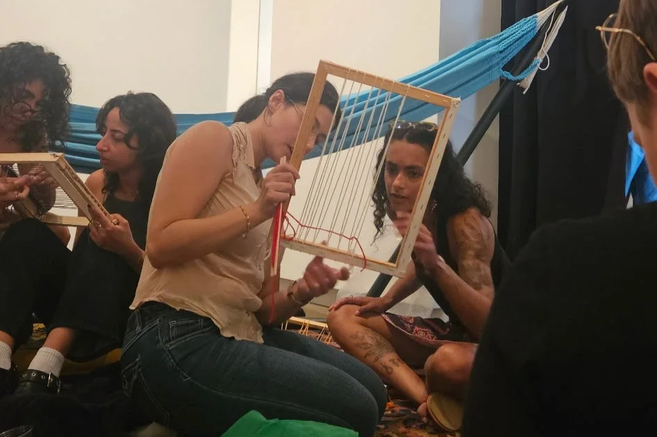 Group of women sitting on the floor, engaged in a craft activity involving wooden frames and strings, with a hammock in the background.