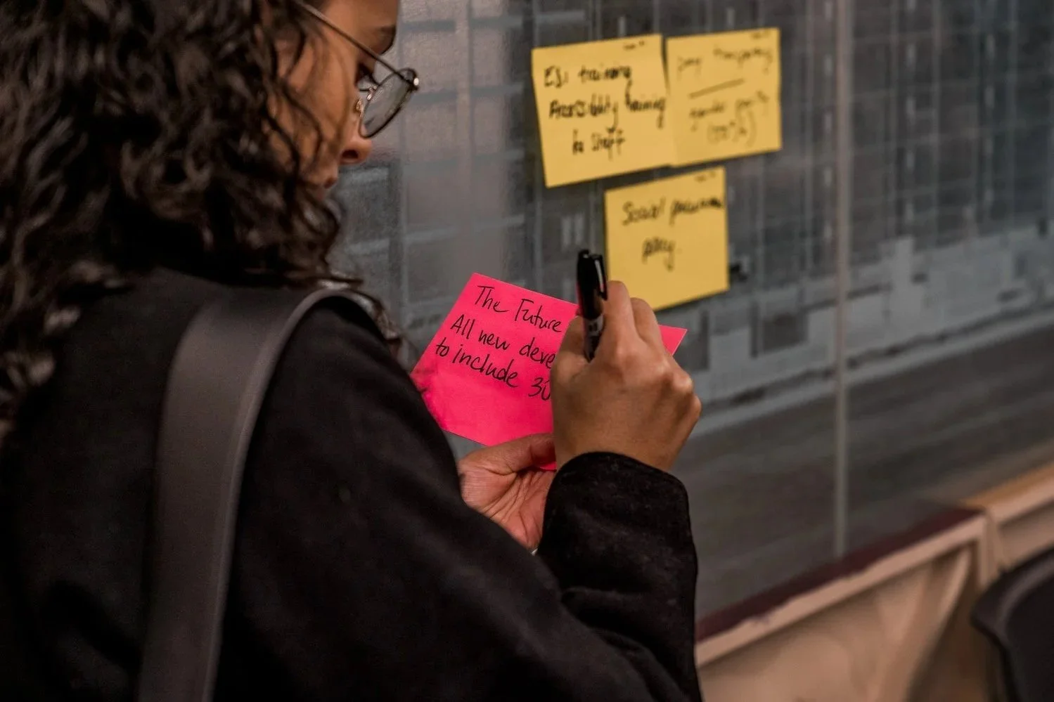 A woman with curly hair and glasses writing on a pink sticky note, with yellow sticky notes on a gray brick wall in the background.