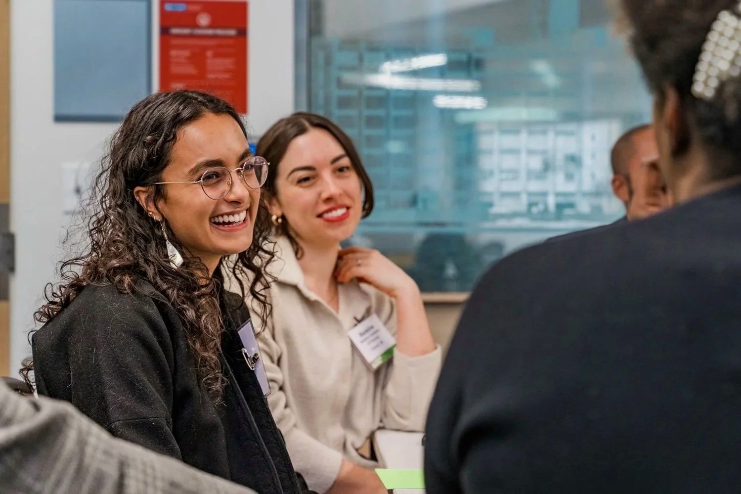 Two women with name tags sitting at a table, smiling and engaged in conversation with others during a professional meeting or networking event.